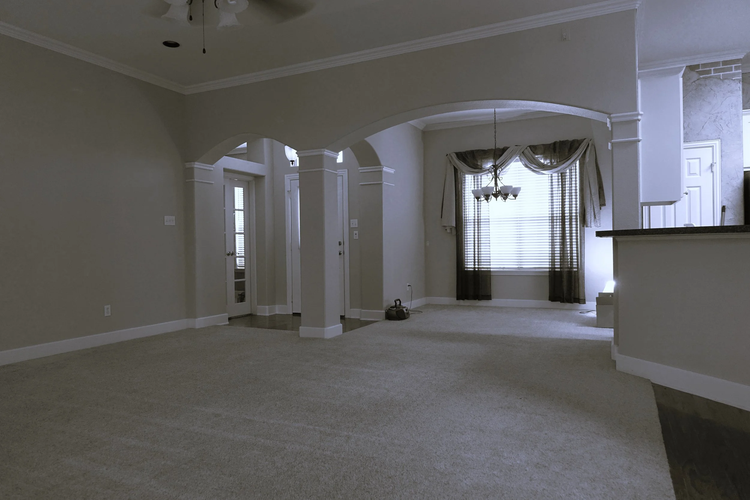 Empty living room with beige walls, carpeted floor, large window with curtains, and a chandelier hanging from the ceiling