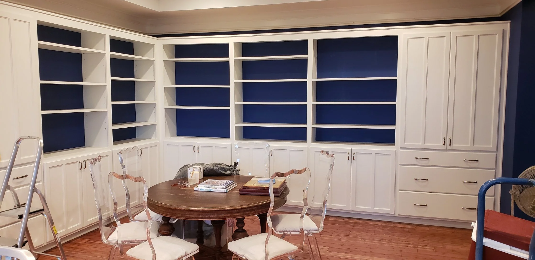 Empty white built-in bookshelf with dark blue backing, surrounding a corner in a room with hardwood flooring, with a round wooden table and six transparent chairs in the center.