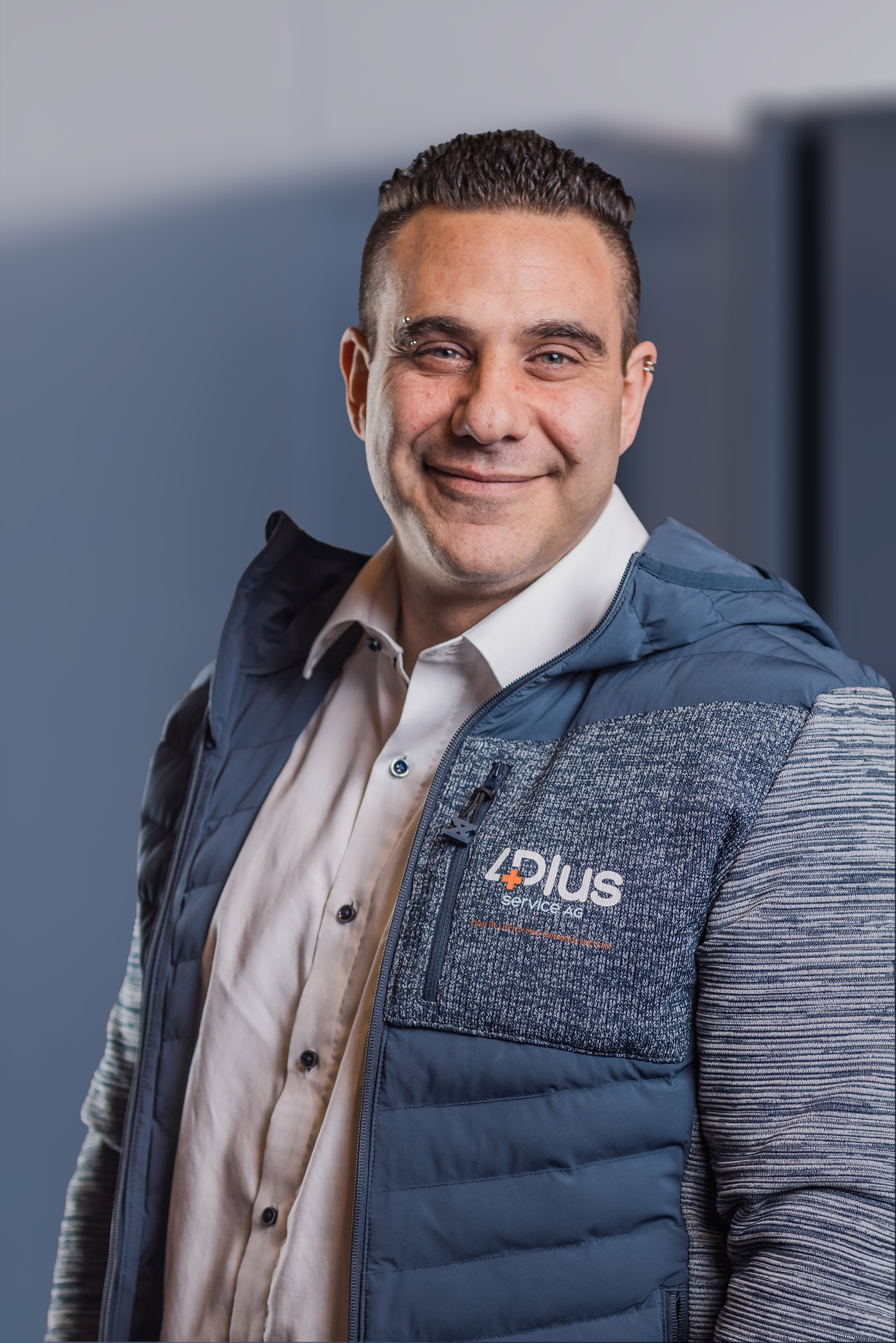 Mann in blau-weißer Jacke mit "4Plus Service AG" Logo, lächelnd, vor neutralem Hintergrund.