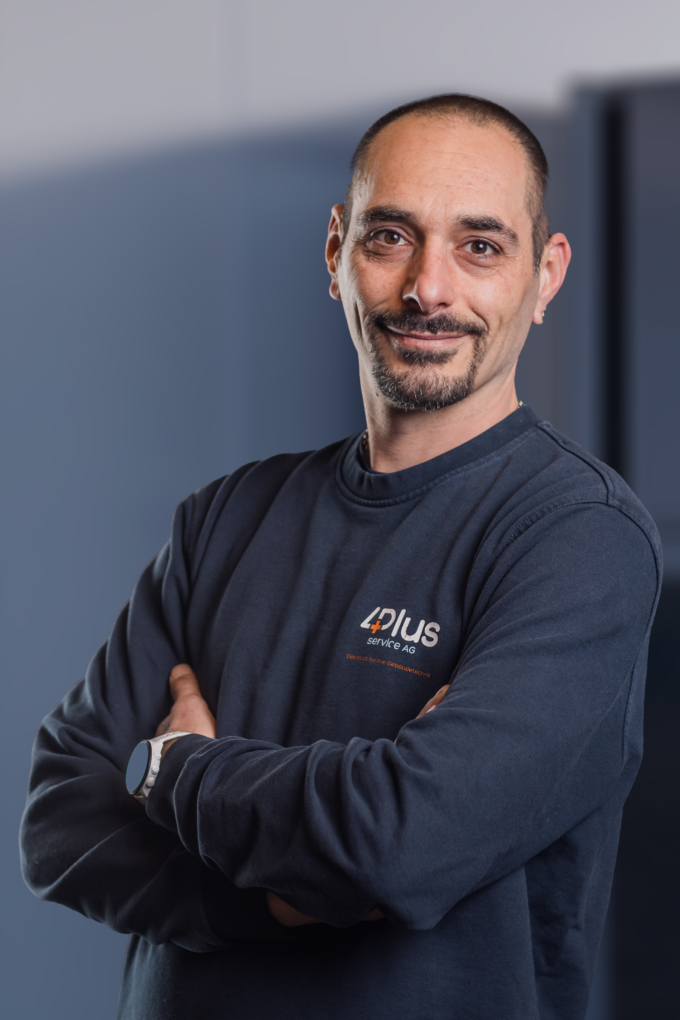 Mann in schwarzem Pullover mit "4plus service AG" Logo, verschränkte Arme, lächelndes Gesicht, Glatze und Bart, neutraler Hintergrund.