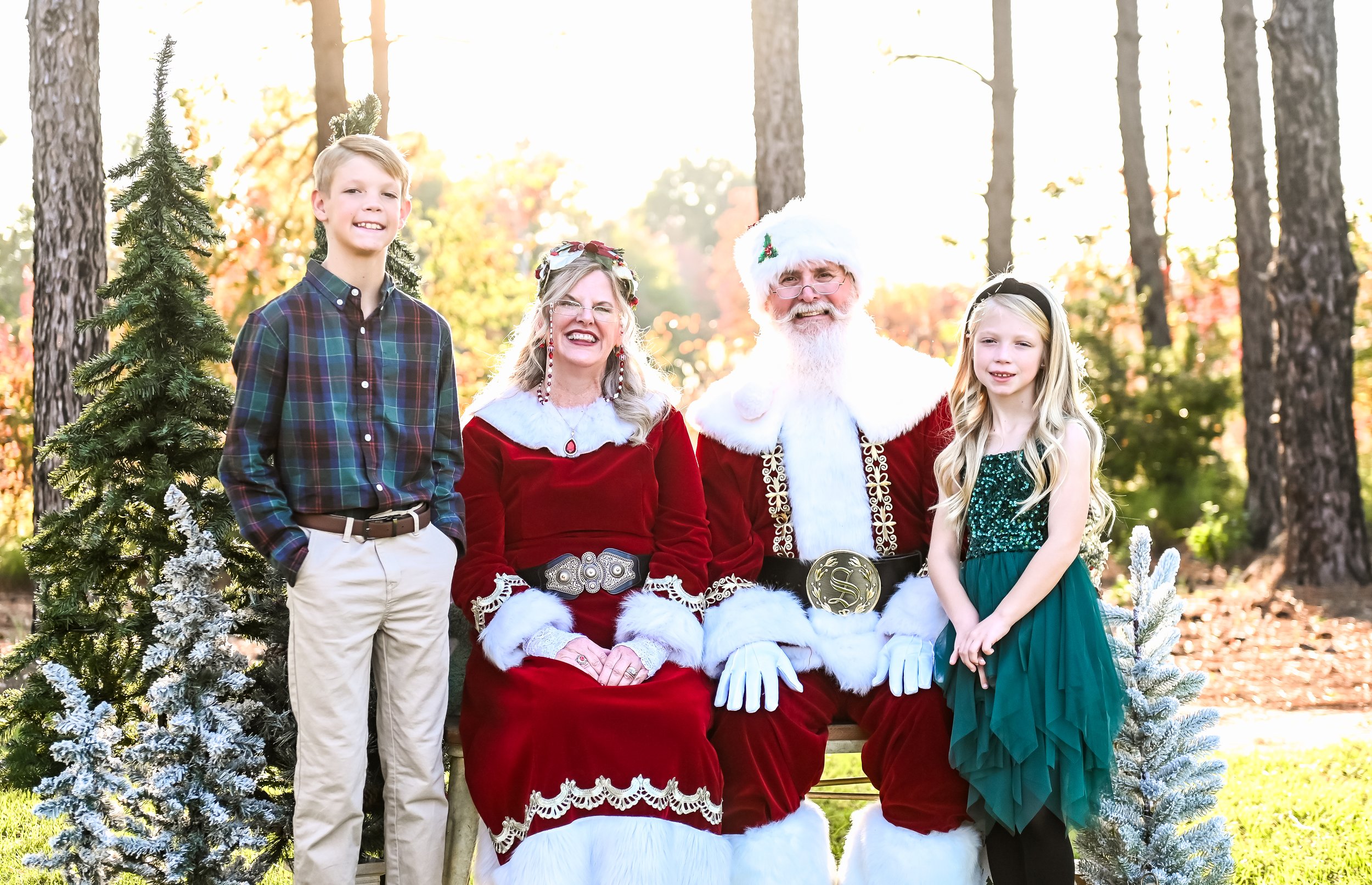 A smiling family photo with Santa and Mrs. Claus, a boy in a plaid shirt, and a girl in a green dress, surrounded by trees and holiday decorations.