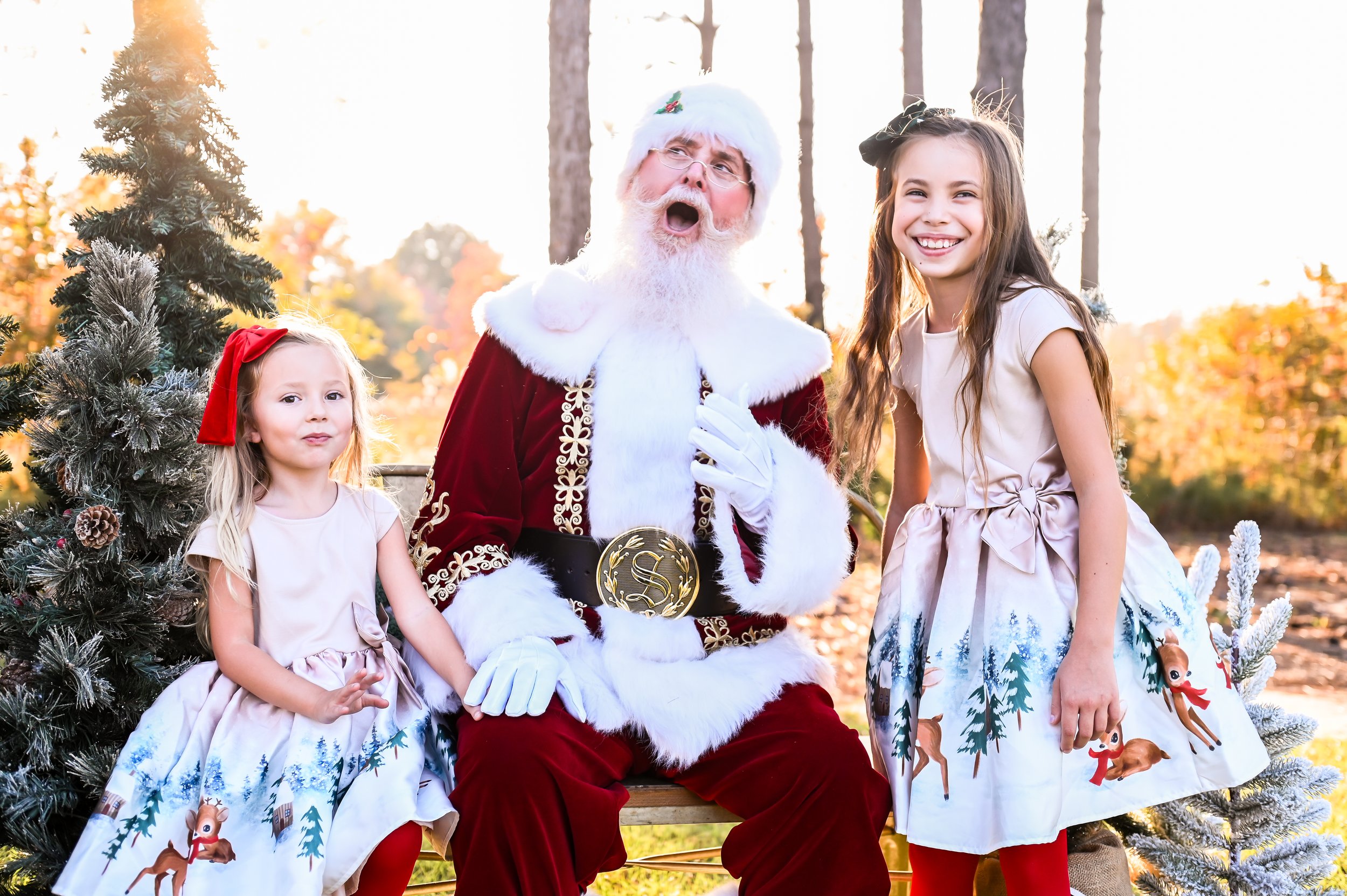 Santa Claus sitting on a bench with two girls in festive dresses, surrounded by pine trees, in a park setting during autumn.