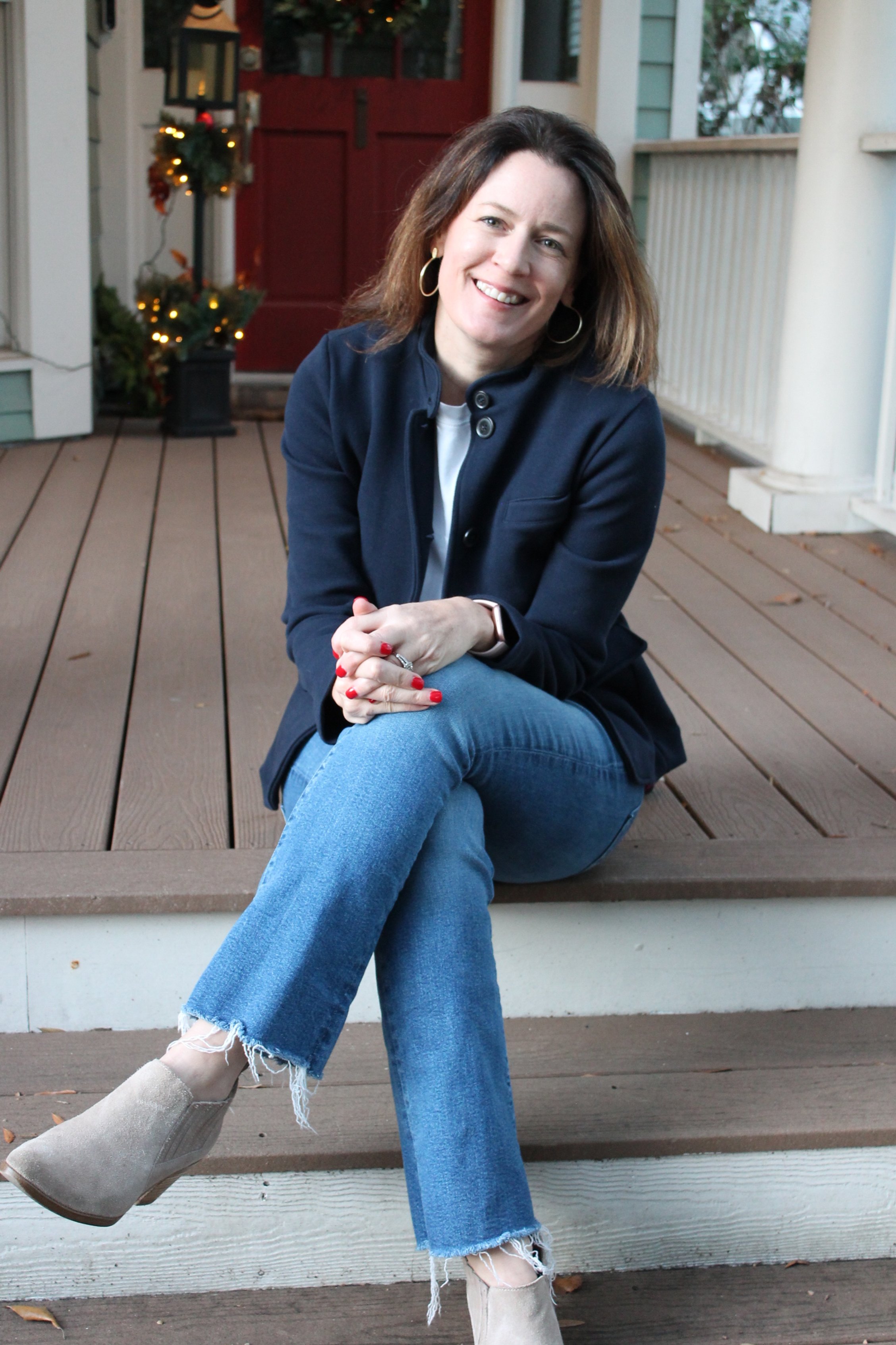 A woman with shoulder-length brown hair, wearing a dark blue jacket, light blue ripped jeans, and beige ankle boots, sitting on the porch of a house with a red door, decorated with Christmas lights and a small wreath.