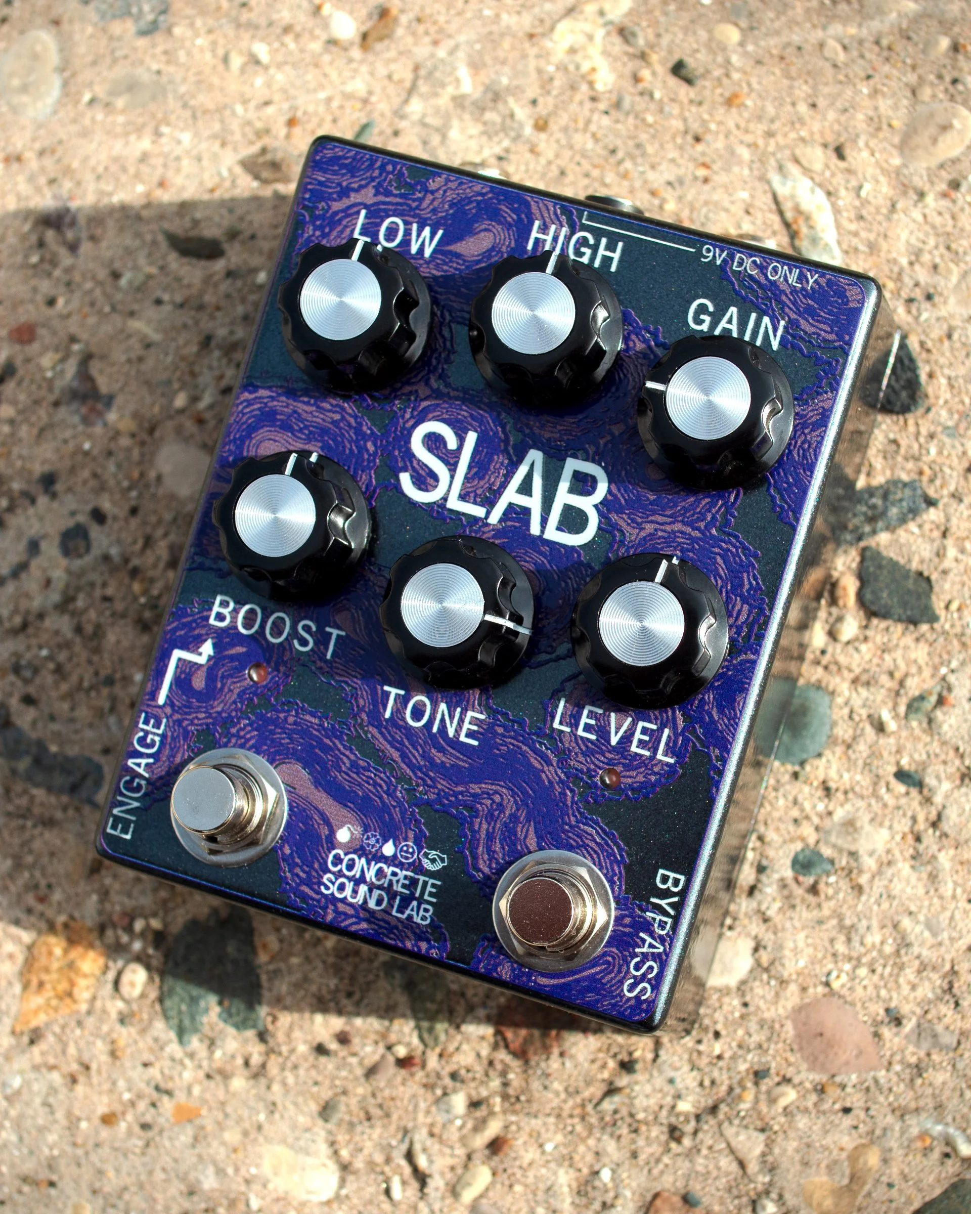 Close-up of a guitar effects pedal with black knobs, set on sandy, rocky ground. The pedal is labeled 'SLAB' and has controls for 'LOW,' 'HIGH,' and 'GAIN,' along with other text and symbols.