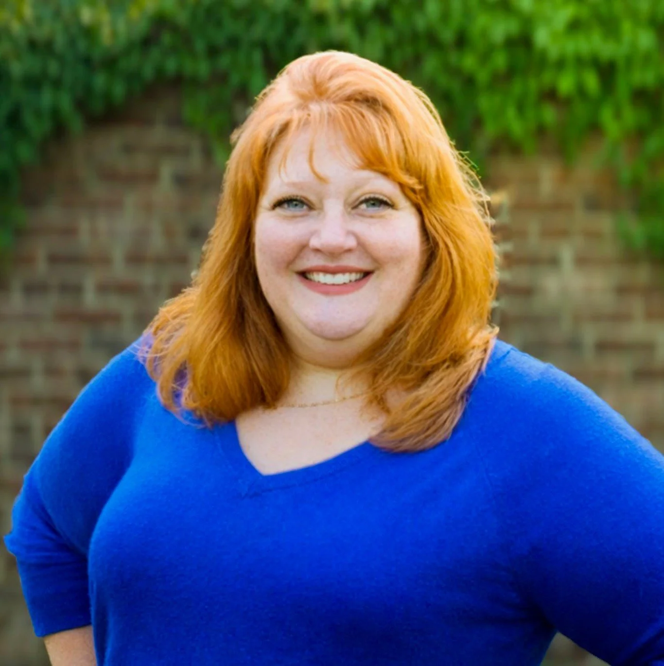 A woman with red hair and a big smile wearing a blue top, standing outdoors with a brick wall and green foliage in the background.