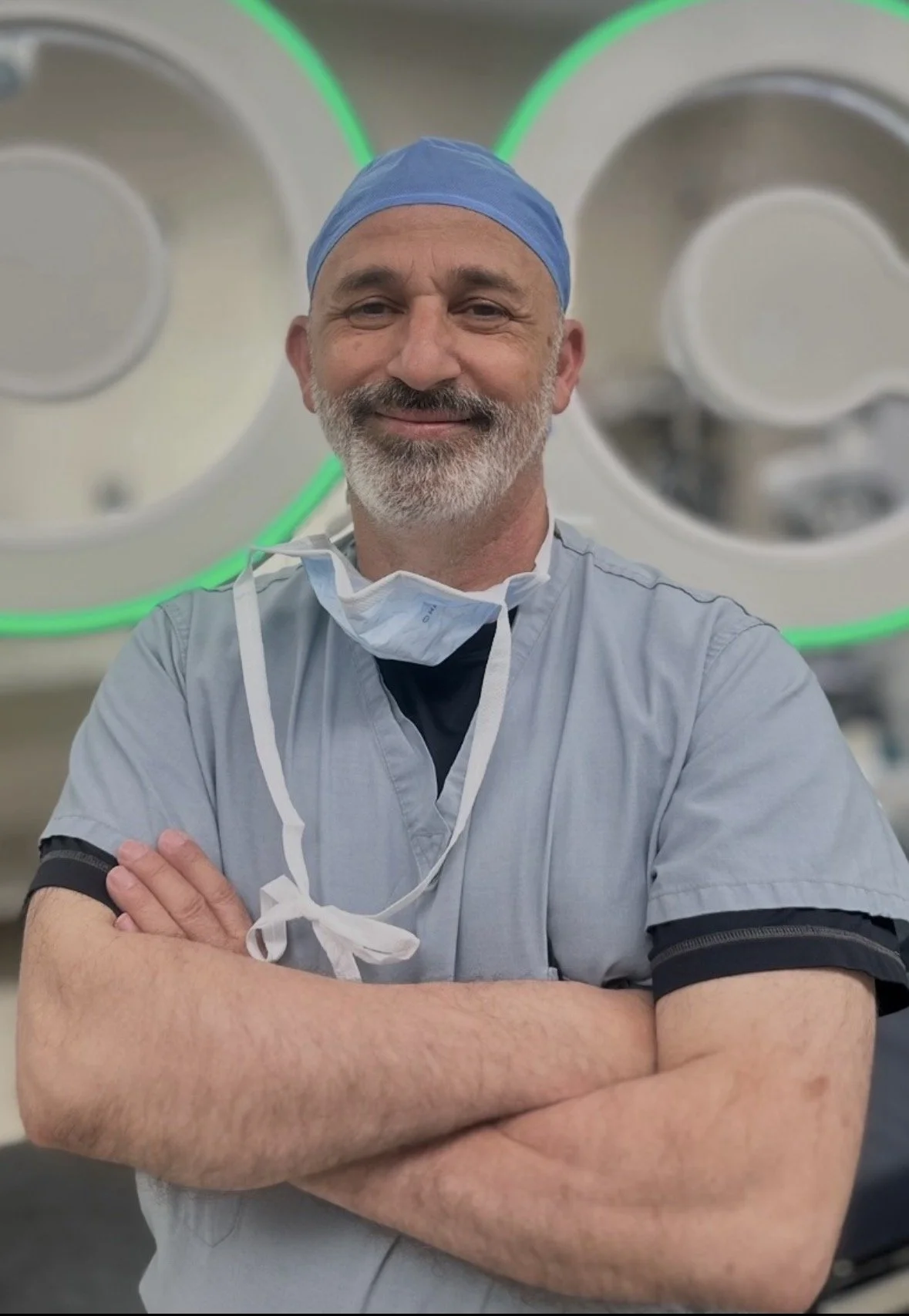 A smiling male surgeon in scrubs and a blue surgical cap standing in an operating room with surgical lights in the background.