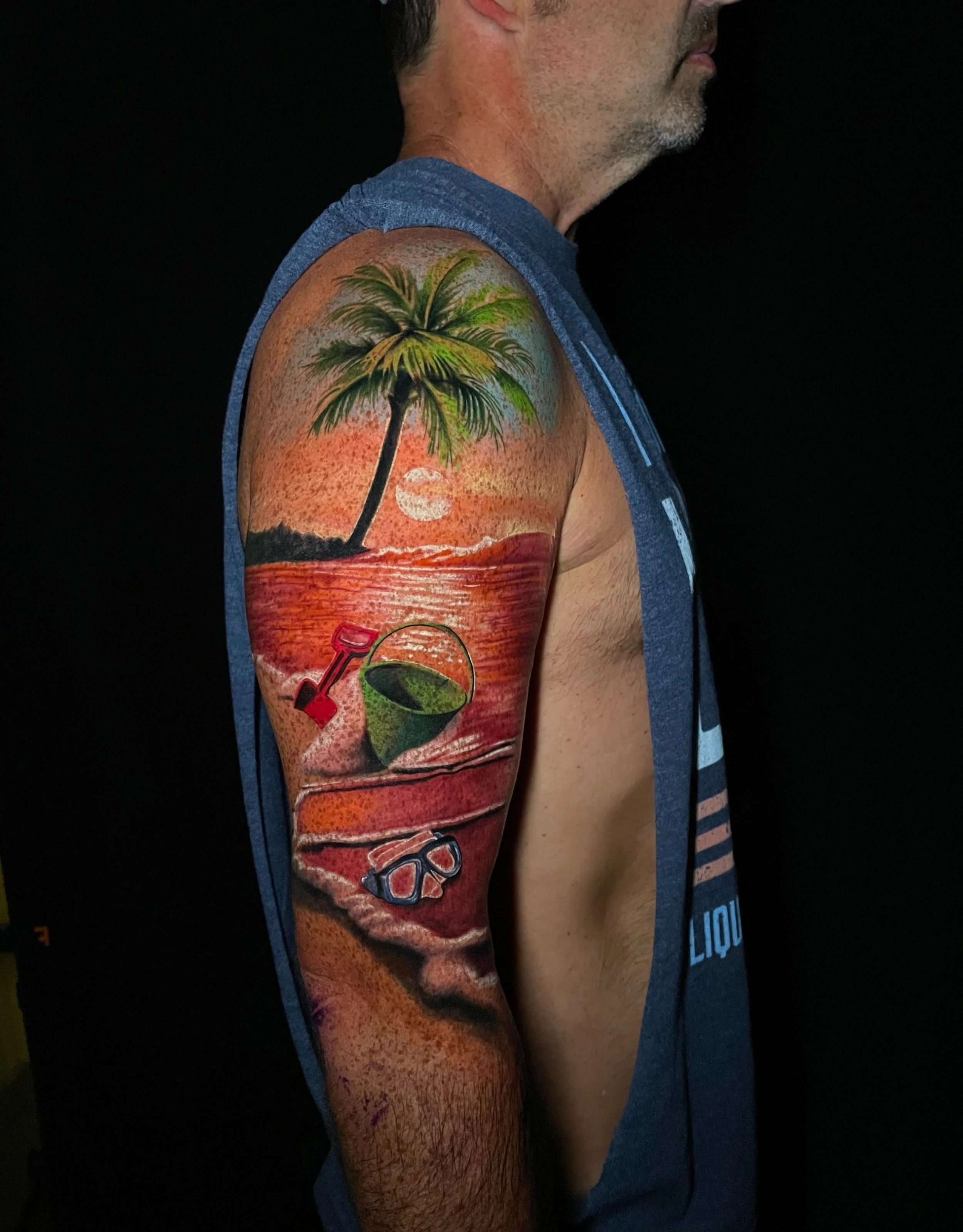 Tattoo of a tropical sunset with a palm tree, a setting sun, a bucket of green beer, a small shovel, and sunglasses on a man's upper arm.