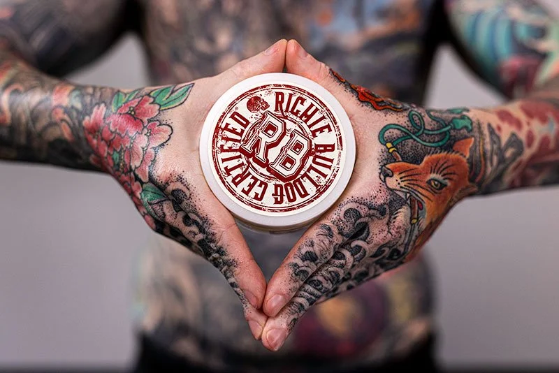 Person with tattoos on their arms and hands holding a container of Riches chocolate chip cookie dough, forming a heart shape with their hands.