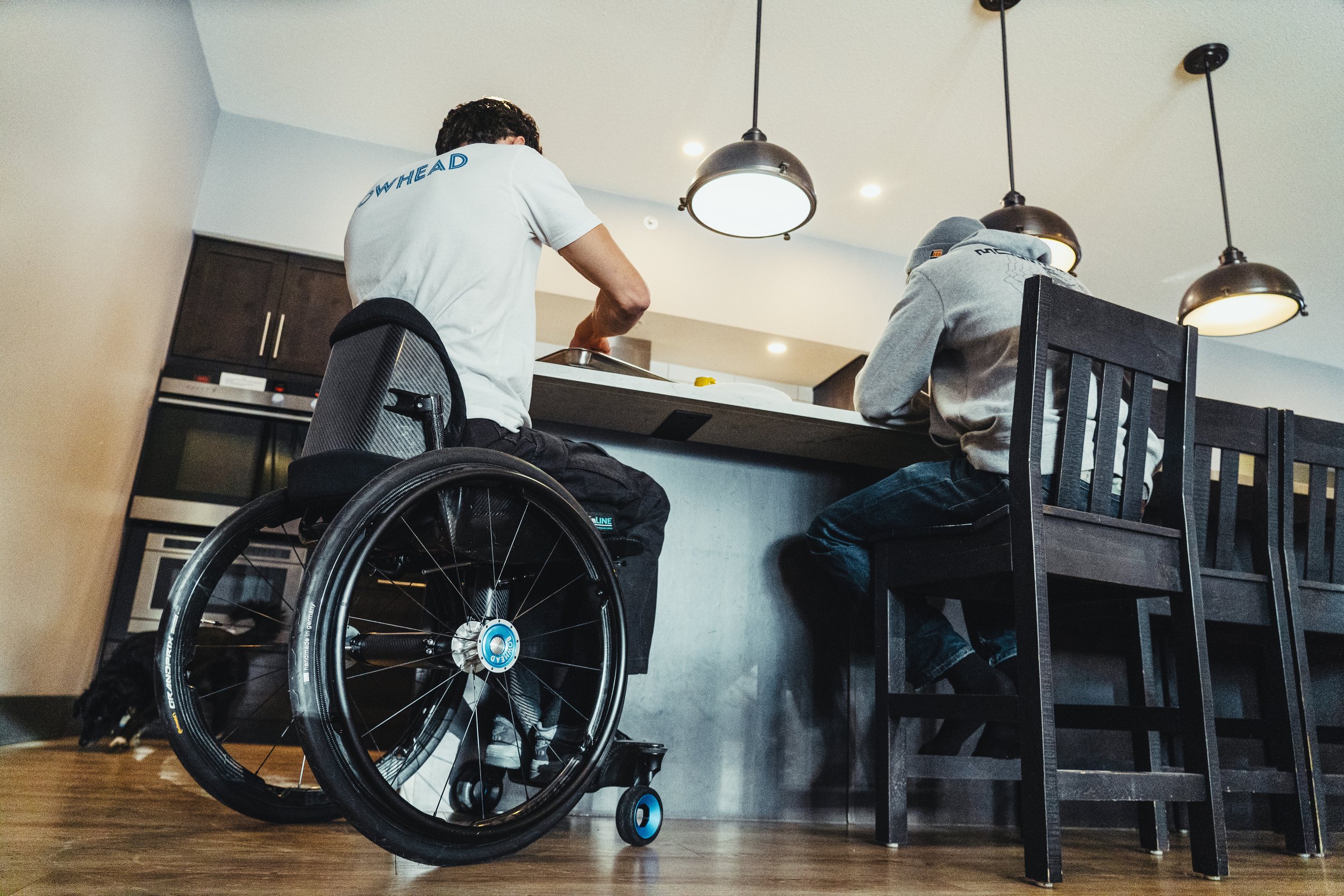 Two people sitting at a kitchen counter, one in a Bowhead Era wheelchair, with modern pendant lights above.