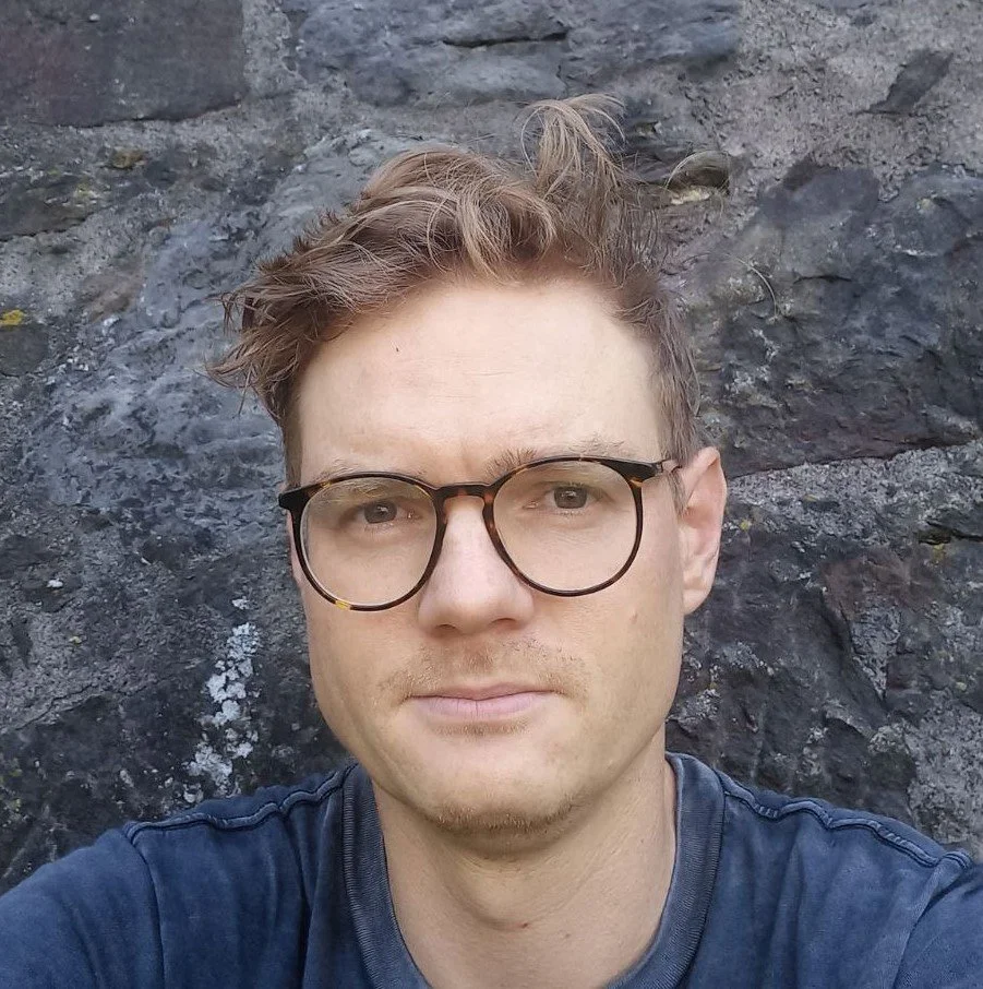 A man with glasses taking a selfie outdoors against a dark rocky background.