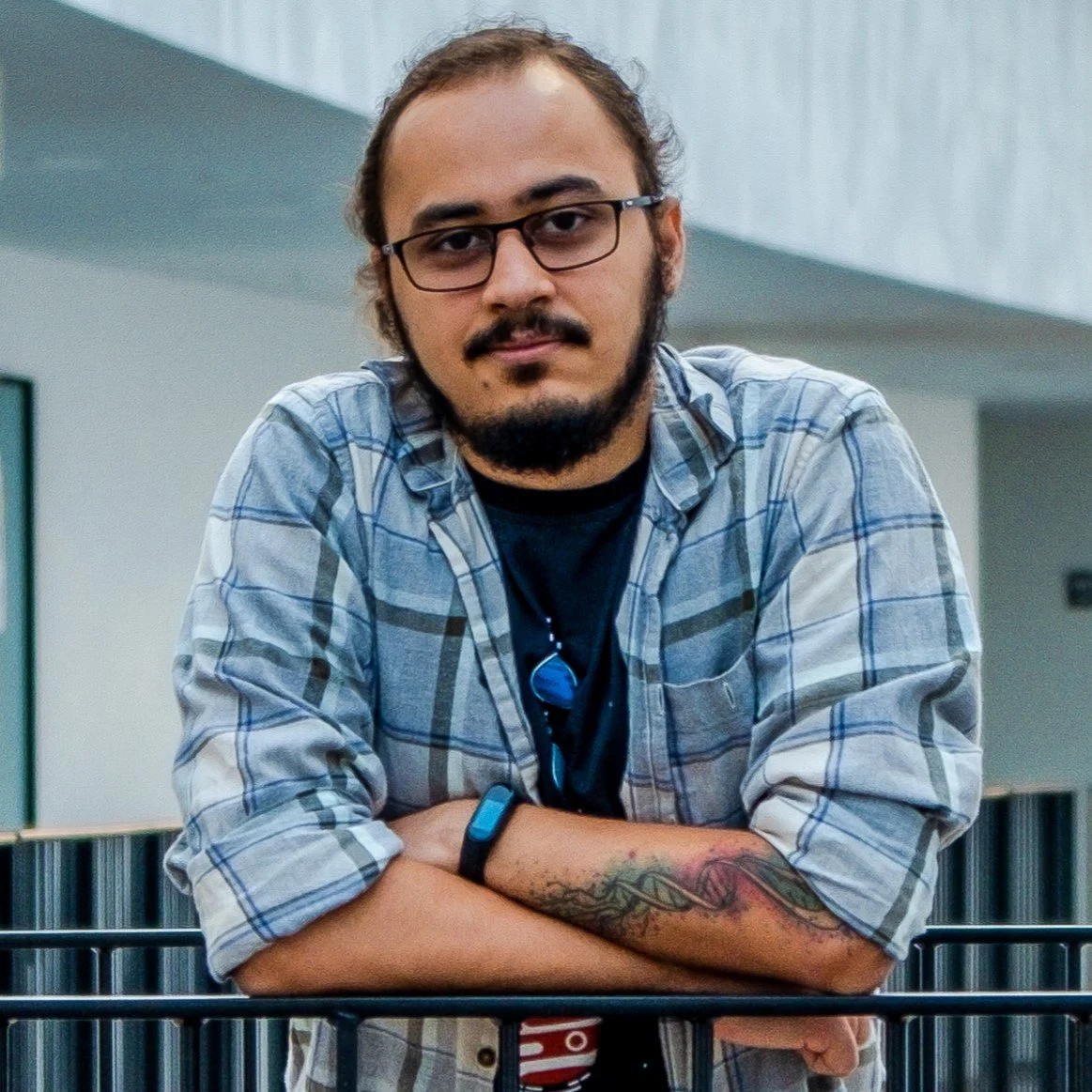 A man with glasses, a beard, and a tattoo on his left arm, wearing a plaid shirt over a black t-shirt, leaning on a black railing in an indoor setting.