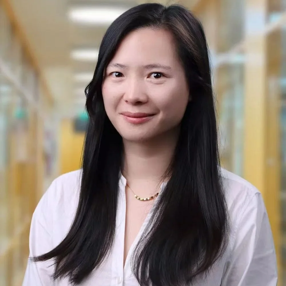 Portrait of an asian woman with brown hair, wearing glasses, a blue collared shirt, and a dark blazer, standing outdoors with blurred building in the background.