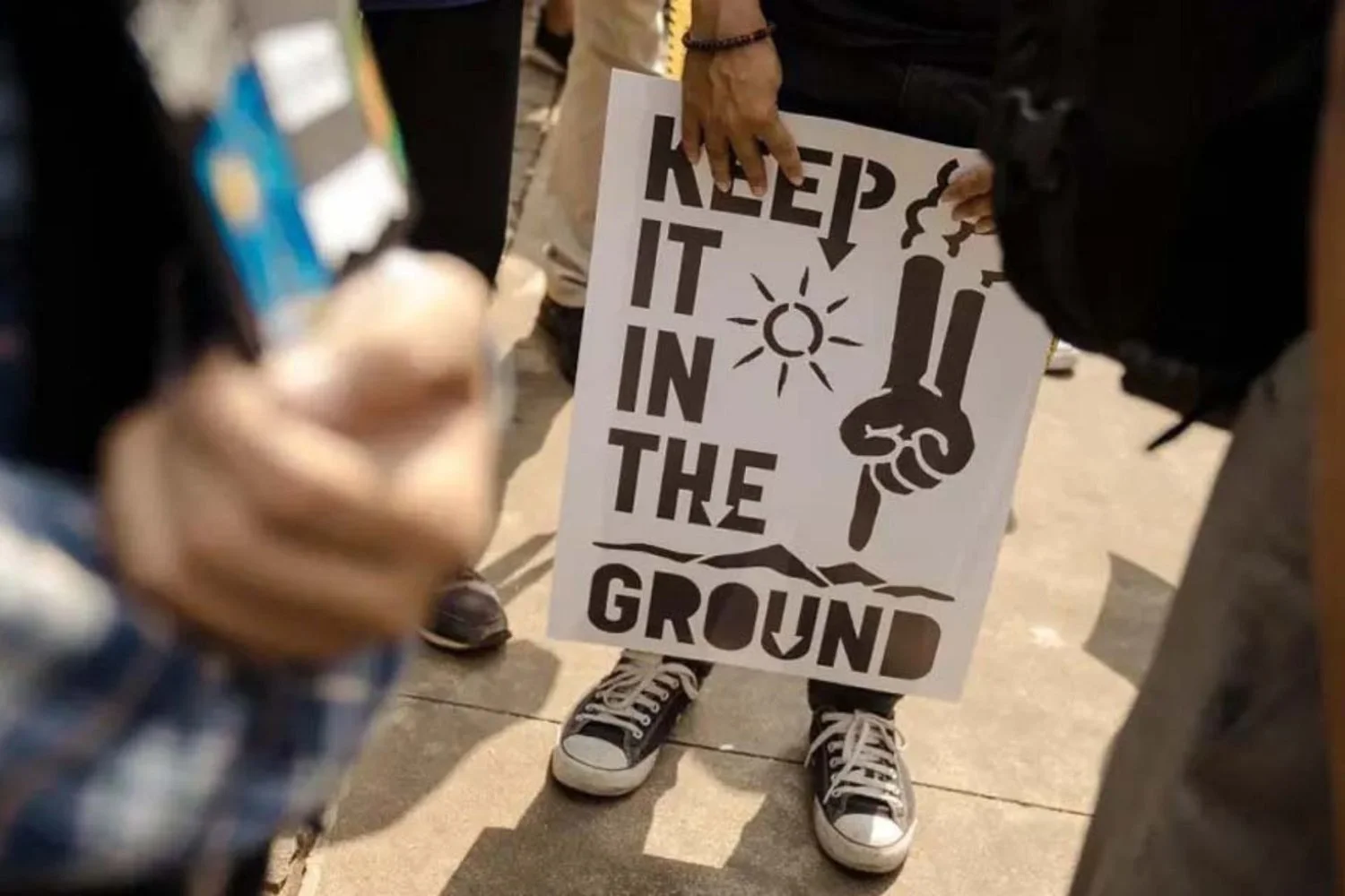 Person holding a protest sign that says "KEEP IT IN THE GROUND" with a sun and pointing hand graphic, standing among a crowd.