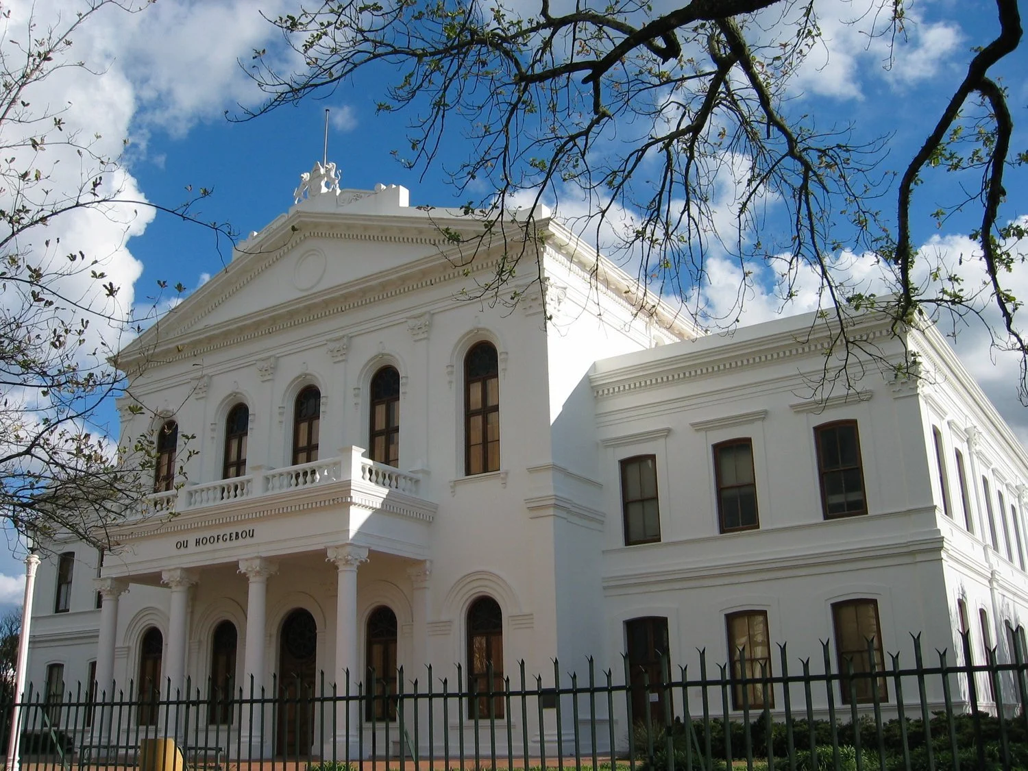 Historic white building with grand columns and large windows behind a black iron fence, under a blue sky with clouds and overhanging tree branches.