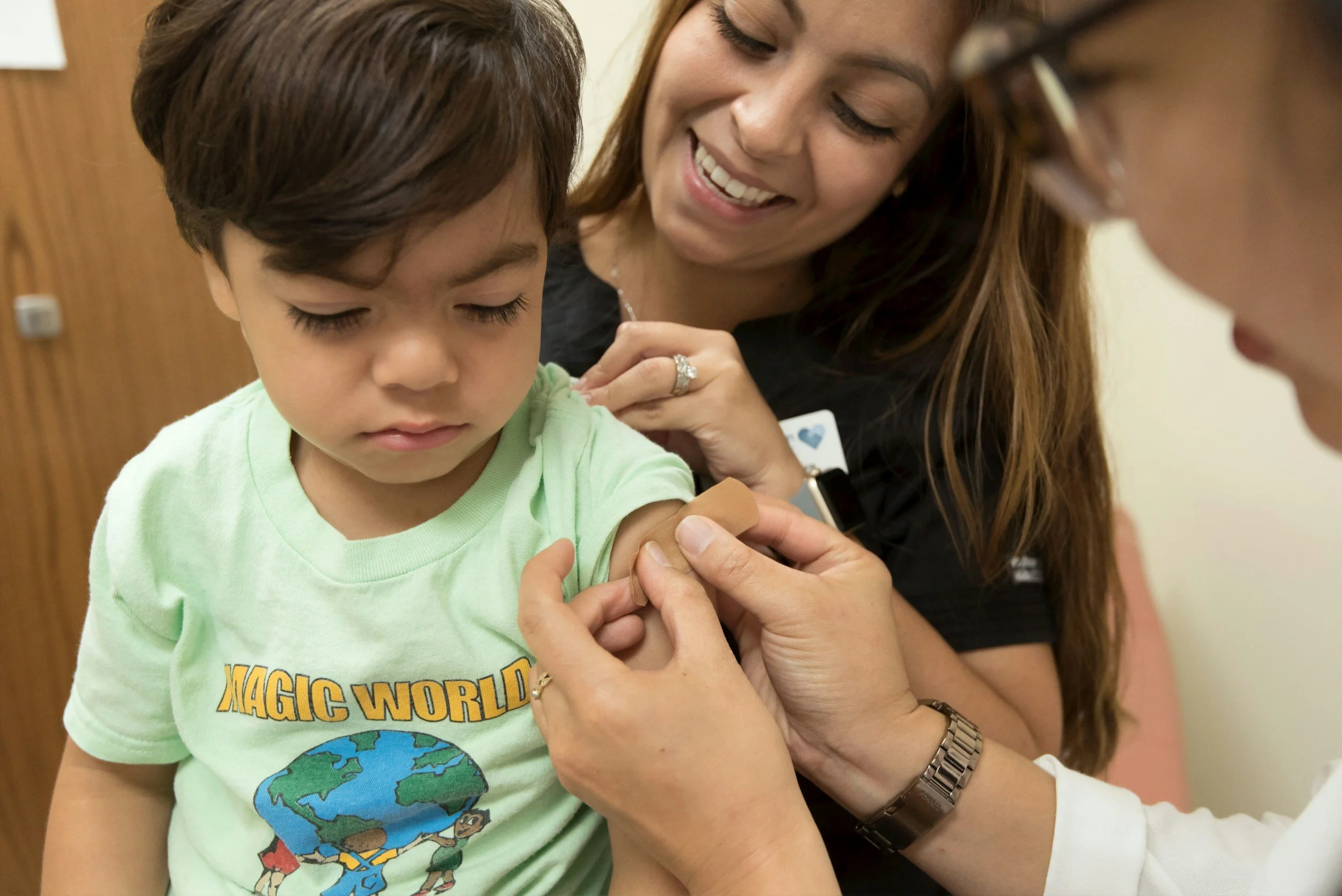 A young boy is receiving a vaccination from a woman, possibly a healthcare professional, who is smiling and looks happy. The boy is wearing a light green t-shirt with a graphic of the Earth and cartoon characters from the movie 'The Magic World.'