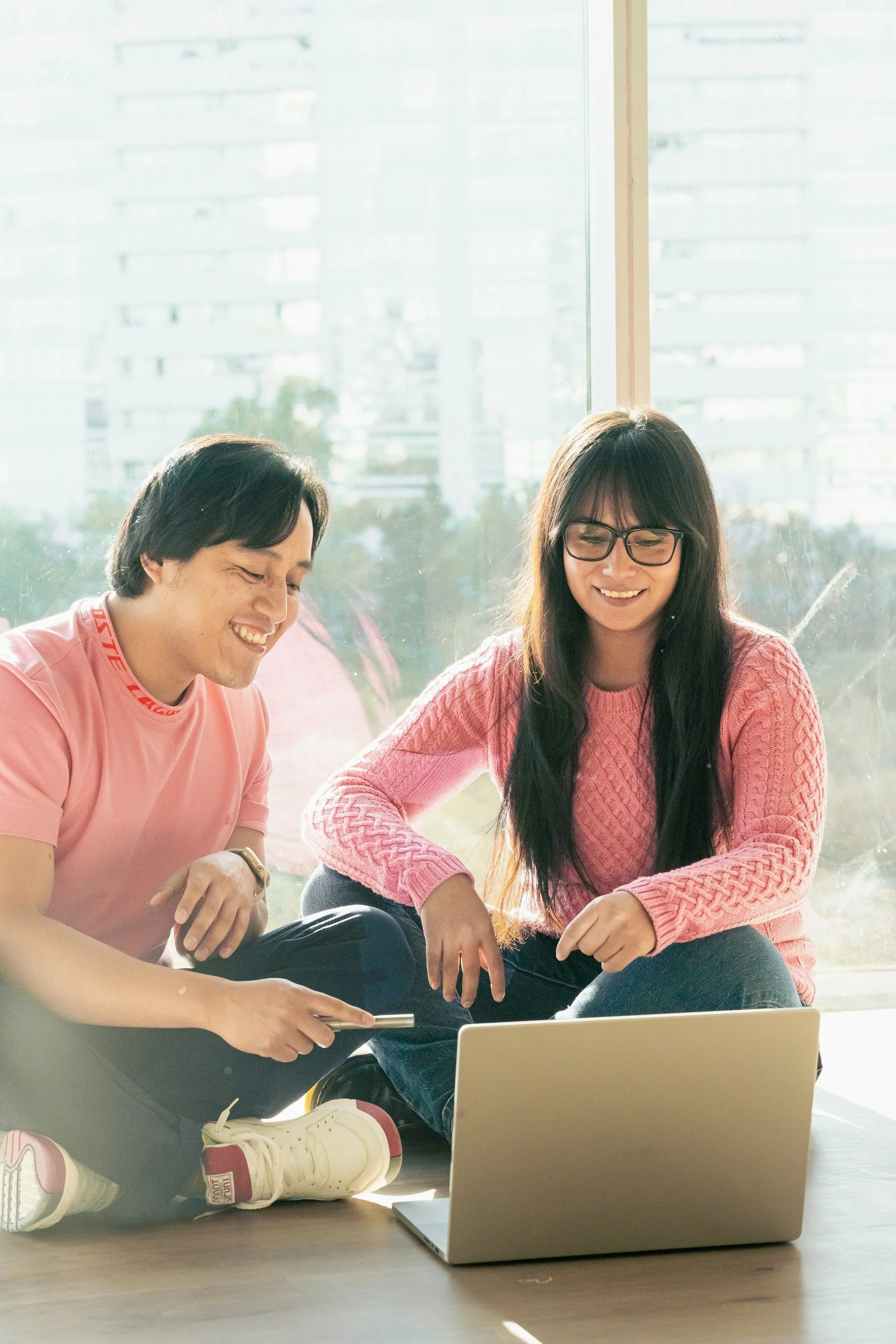 Two people sitting on the floor near a window, looking at a laptop screen, smiling, wearing casual pink clothes, with a laptop in front of them.