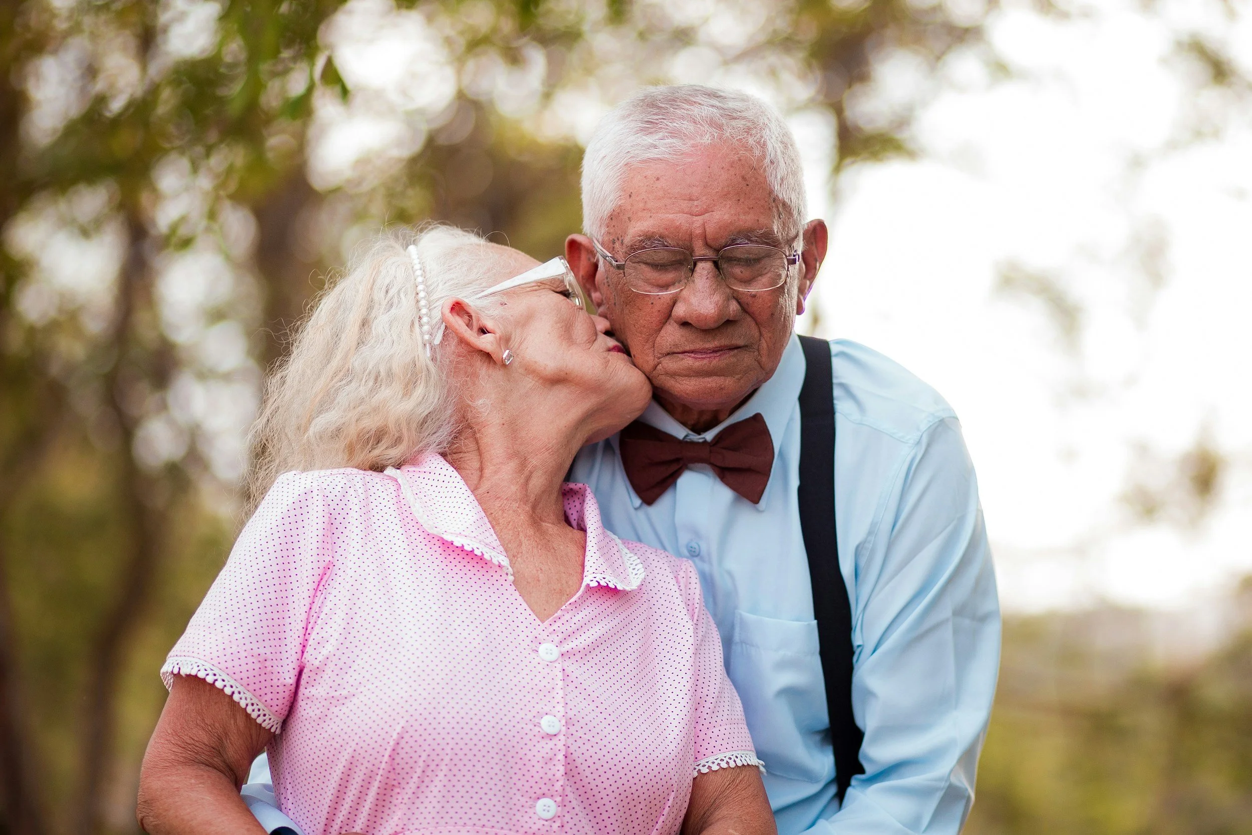 An elderly man with gray hair, glasses, and a bowtie hugs an elderly woman with white hair, glasses, earrings, and a pink polka dot shirt outdoors among trees.