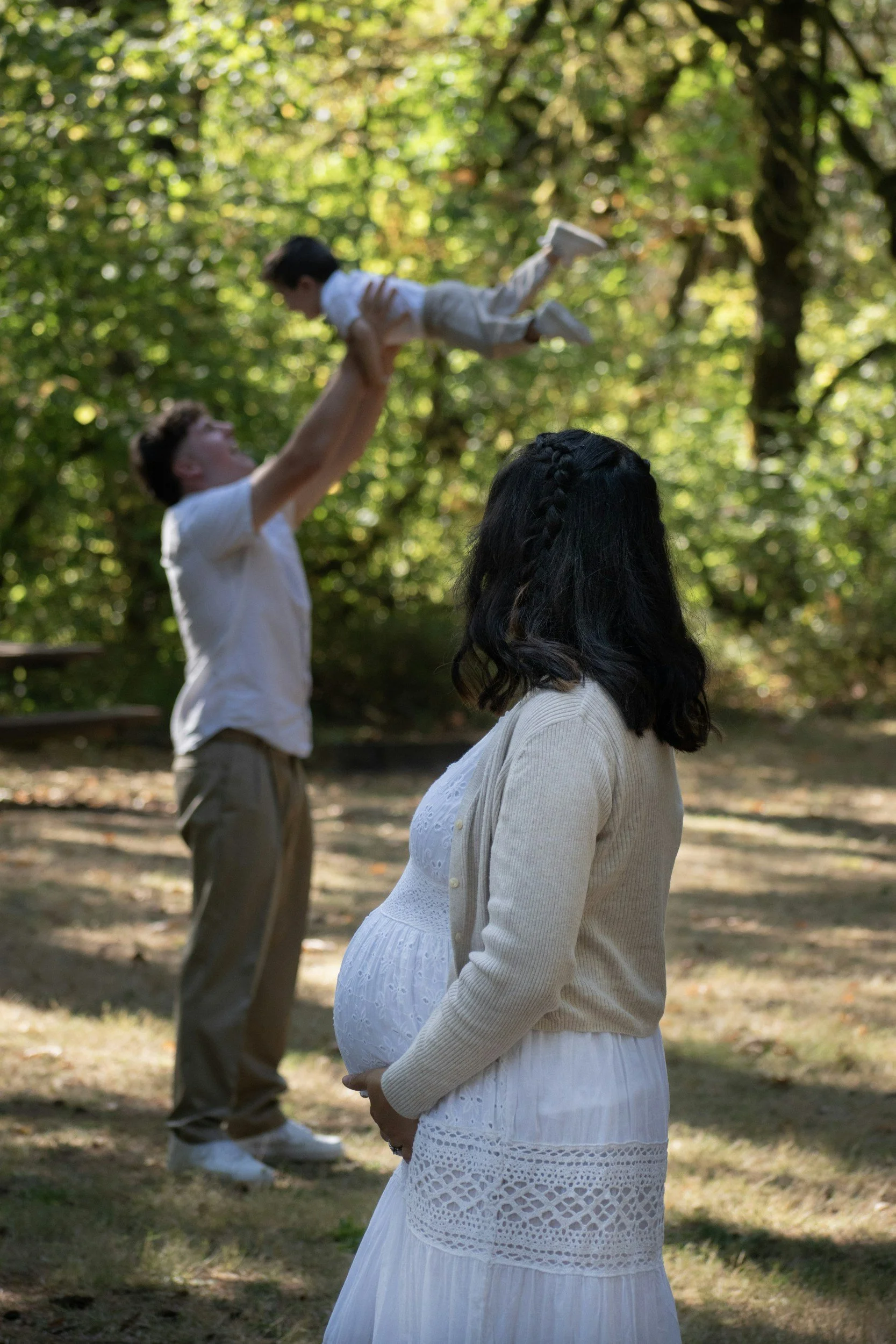 A man lifting a young boy in the air outdoors among trees, with a woman in a white dress and light sweater watching in the foreground.