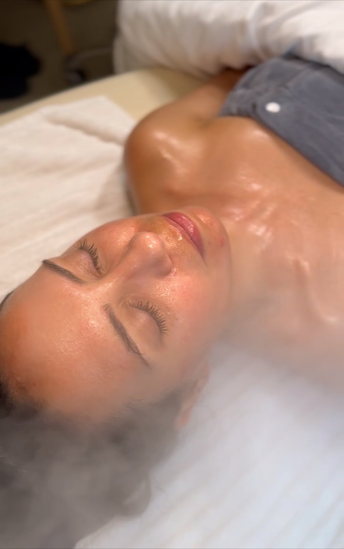 A woman lying on her back with her eyes closed, receiving a facial treatment in a spa or clinic setting.