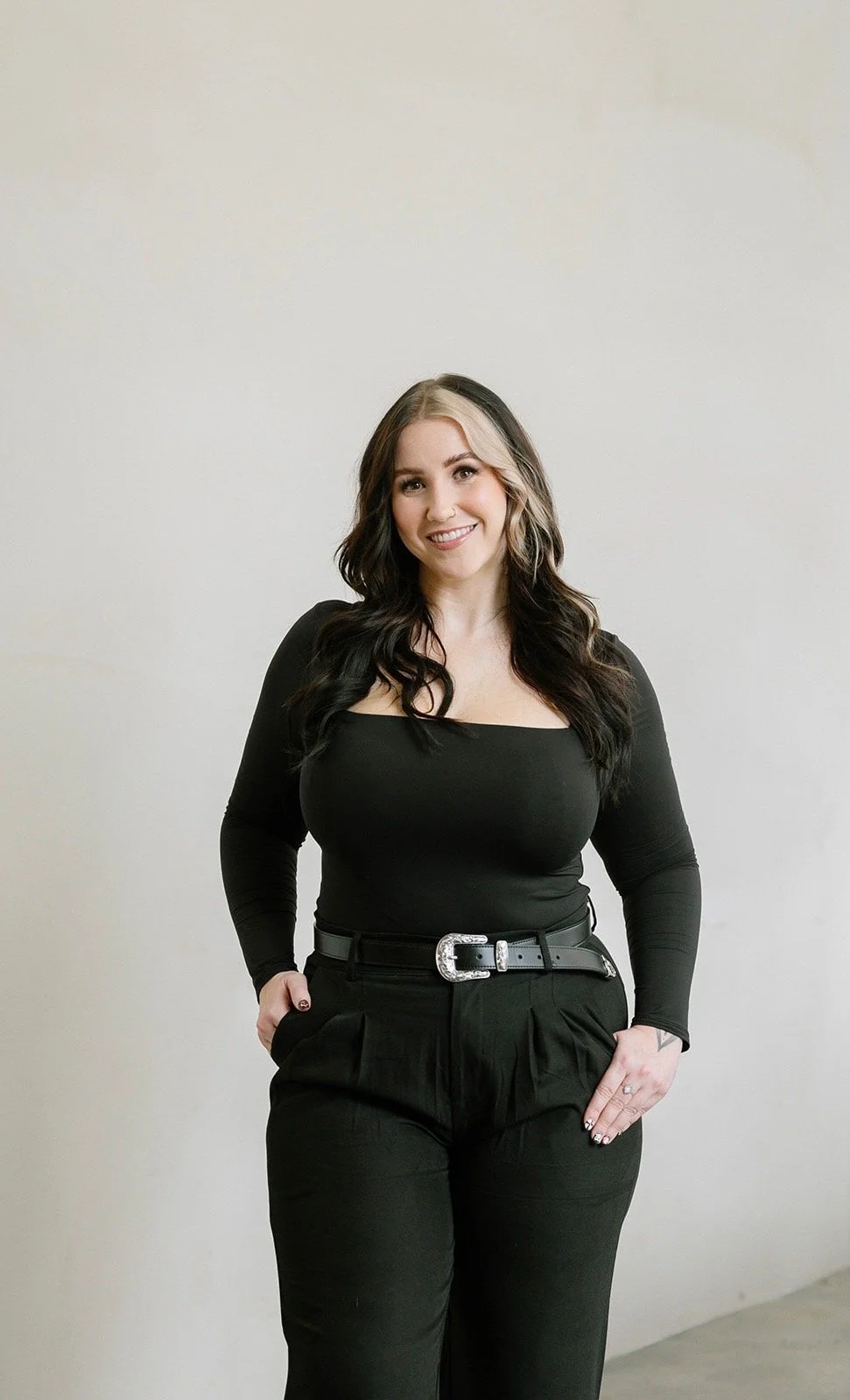 A woman with dark hair and light highlights, wearing a black long-sleeve top, black pants, and a black belt with a silver buckle, stands against a plain off-white background, smiling at the camera.
