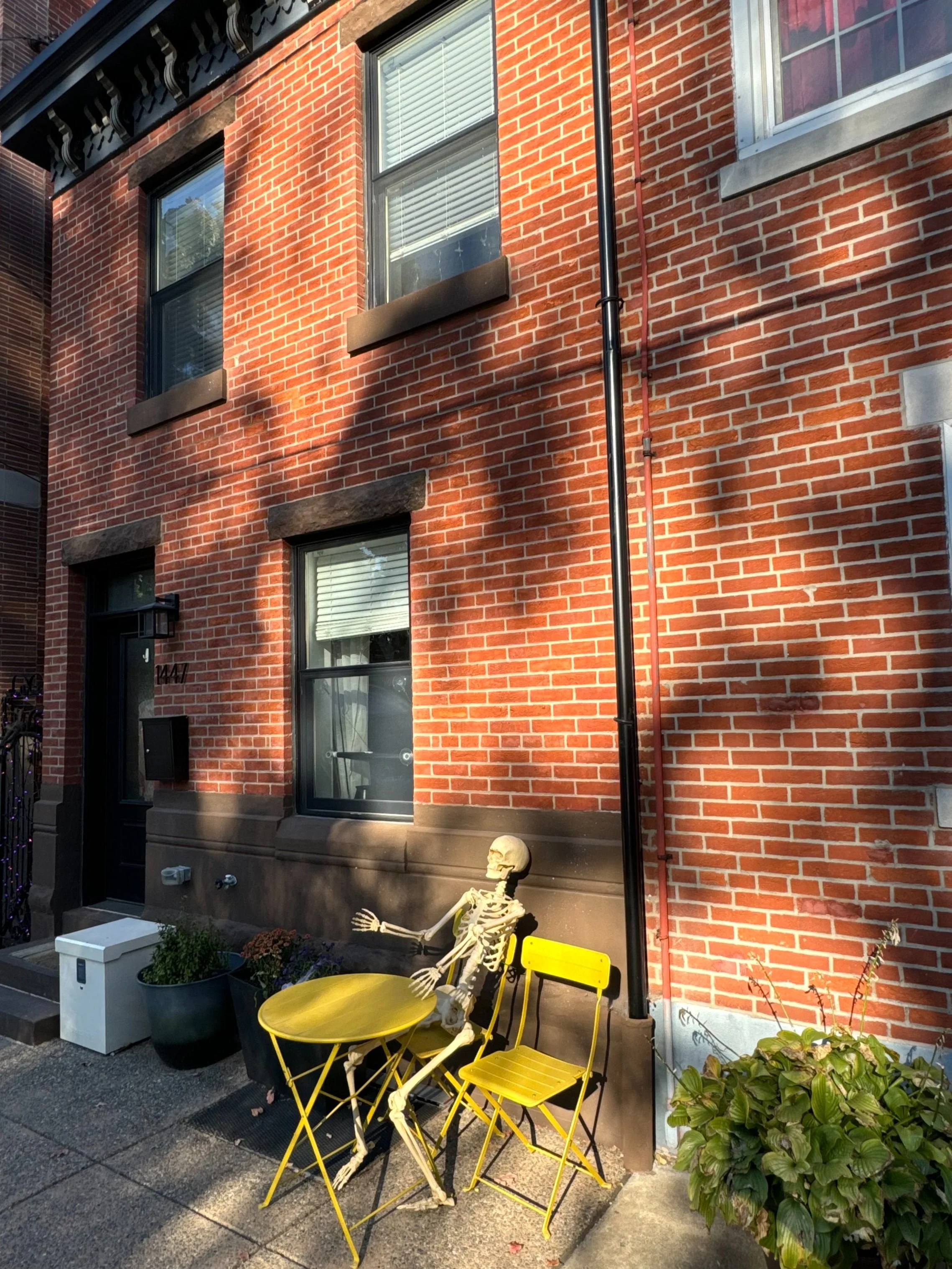 Skeleton sitting outside on a yellow chair, reaching out with its left arm, next to a yellow table and another yellow chair, in front of a brick building with three windows and a black door.