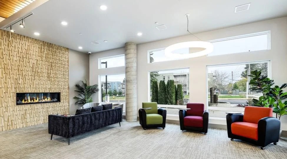 Amenity lounge area featuring a linear fireplace, seating areas, large windows, and indoor landscaping.