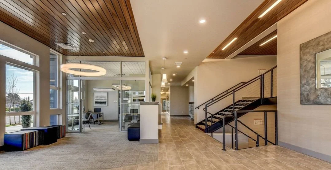 Lobby showing seating areas, glass-enclosed office space, and an open stairwell with wood ceiling accents.