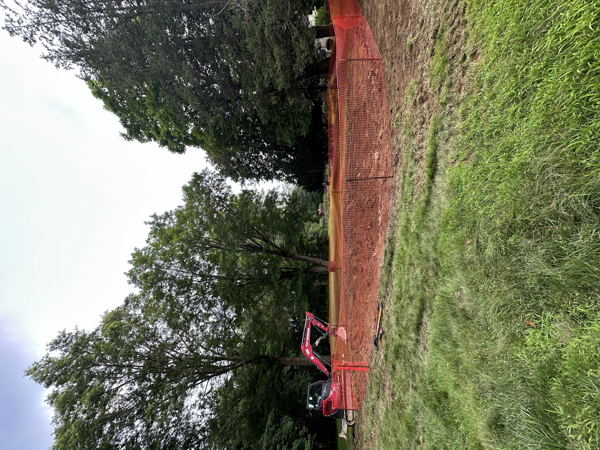 A construction site with an orange safety fence surrounding a trench or excavation area, with a red mini excavator parked on the grass nearby, and trees in the background.