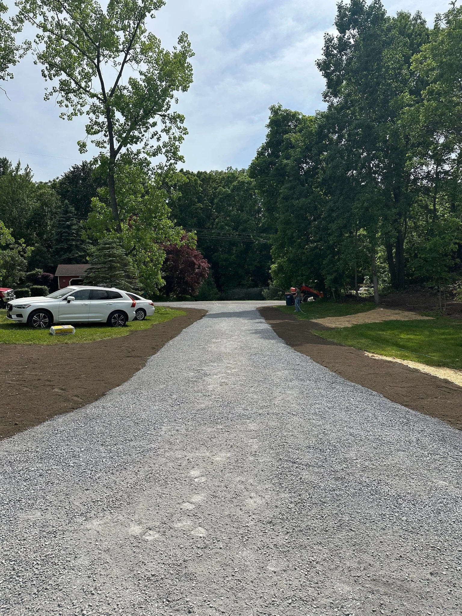 A gravel driveway leading to a residential area with parked cars and lush green trees.