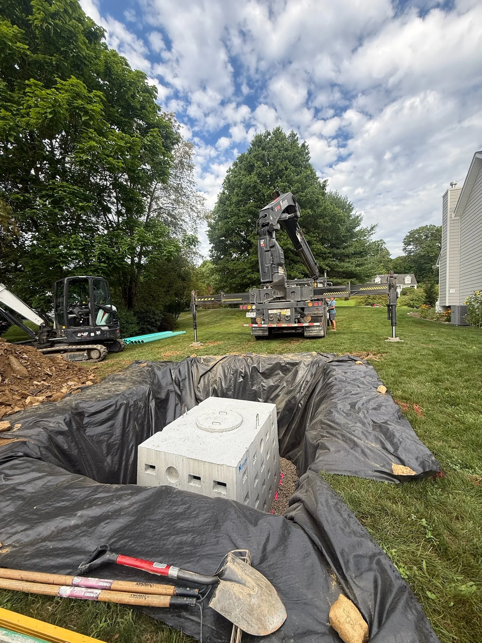 Construction site with a large truck and crane, concrete block, shovels, and excavation equipment on a grassy area under a partly cloudy sky.
