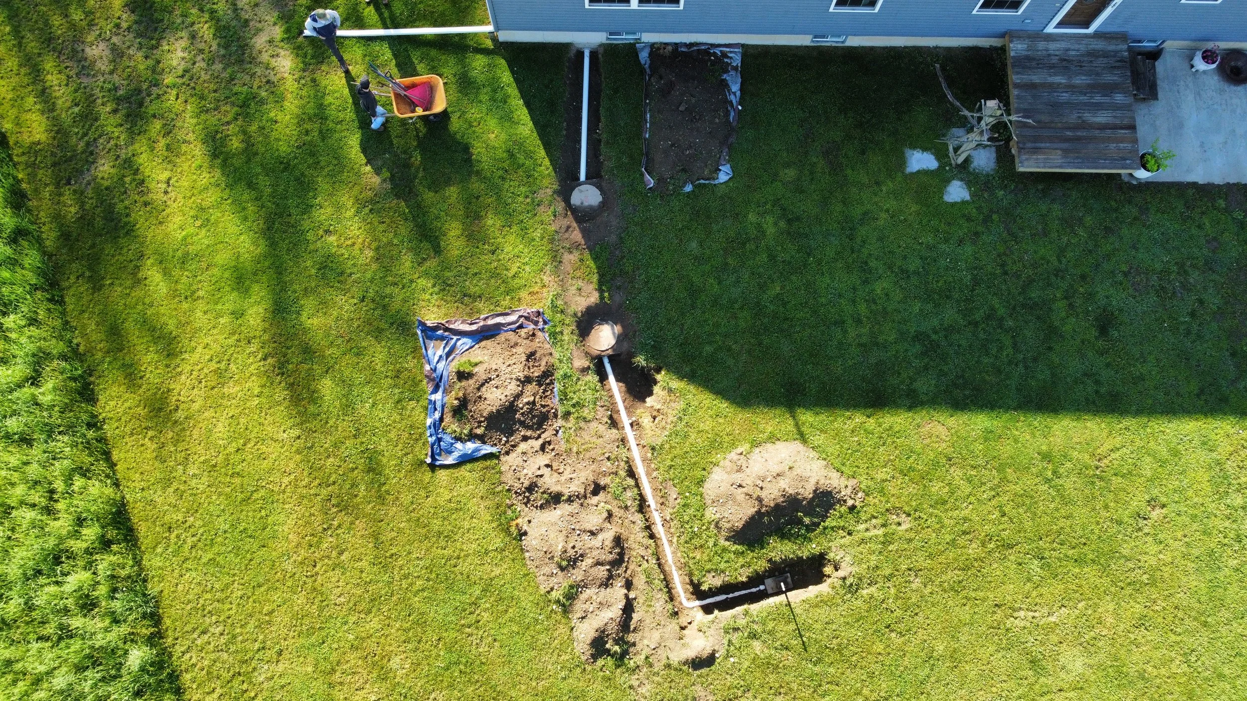An aerial view of a backyard showing a trench being dug by workers for installing underground pipes near a house with a grassy lawn and a deck.