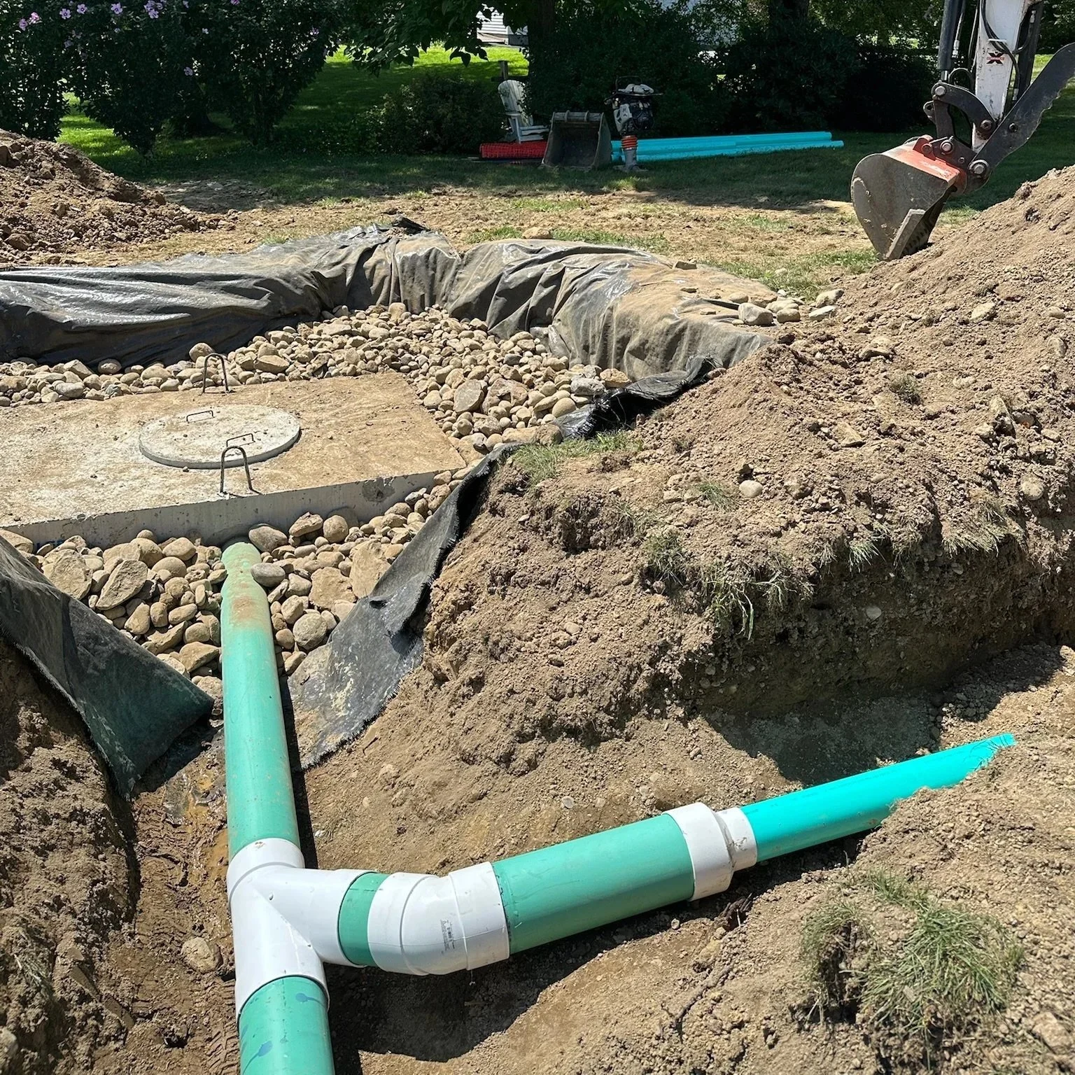 Underground plumbing pipes in a yard under construction next to a trench filled with rocks and dirt, with a large tree and garden tools in the background.
