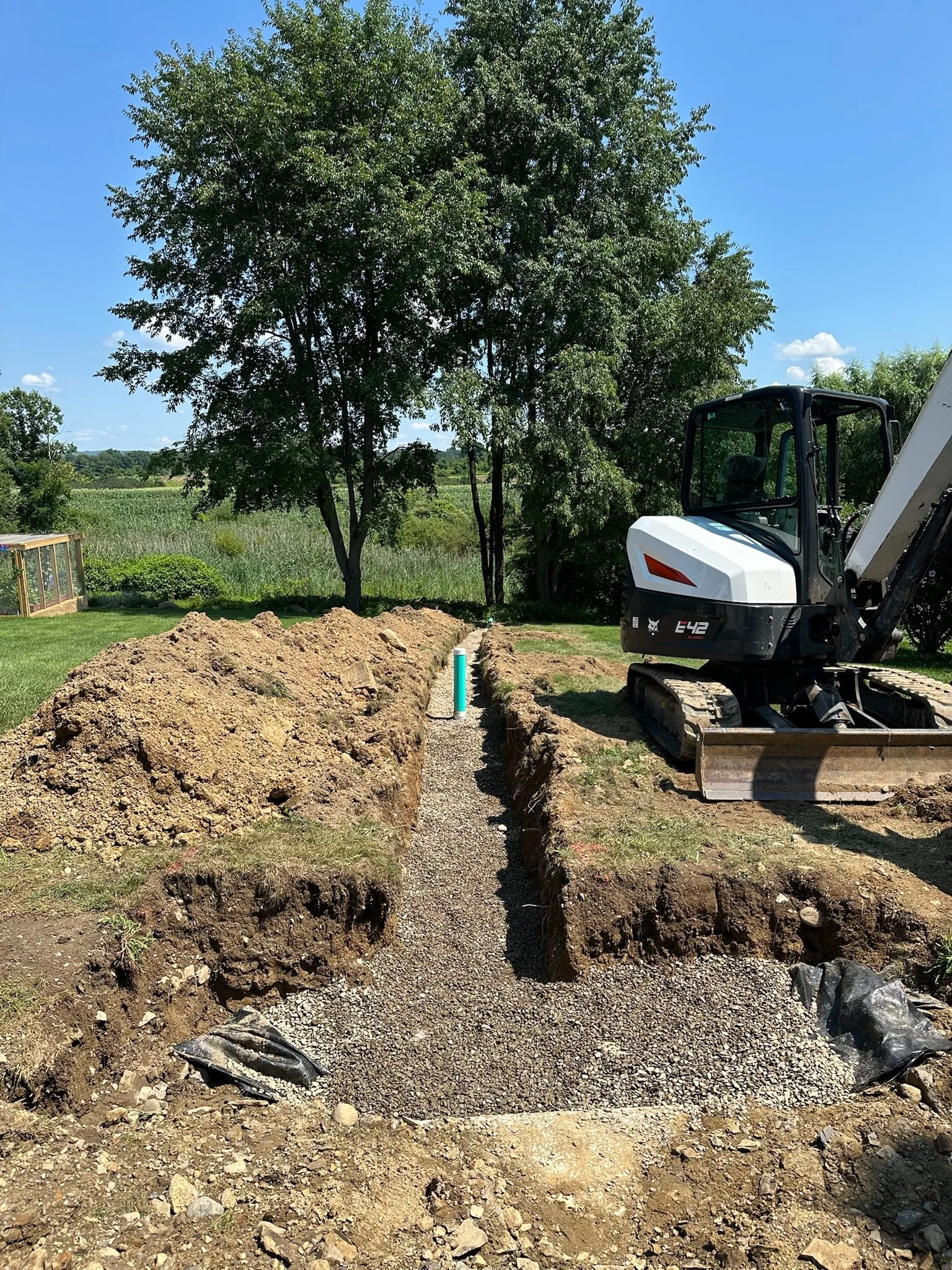 Excavation site with a small trench, dirt pile, and construction equipment on a grassy field, trees, blue sky, and a wooded background.