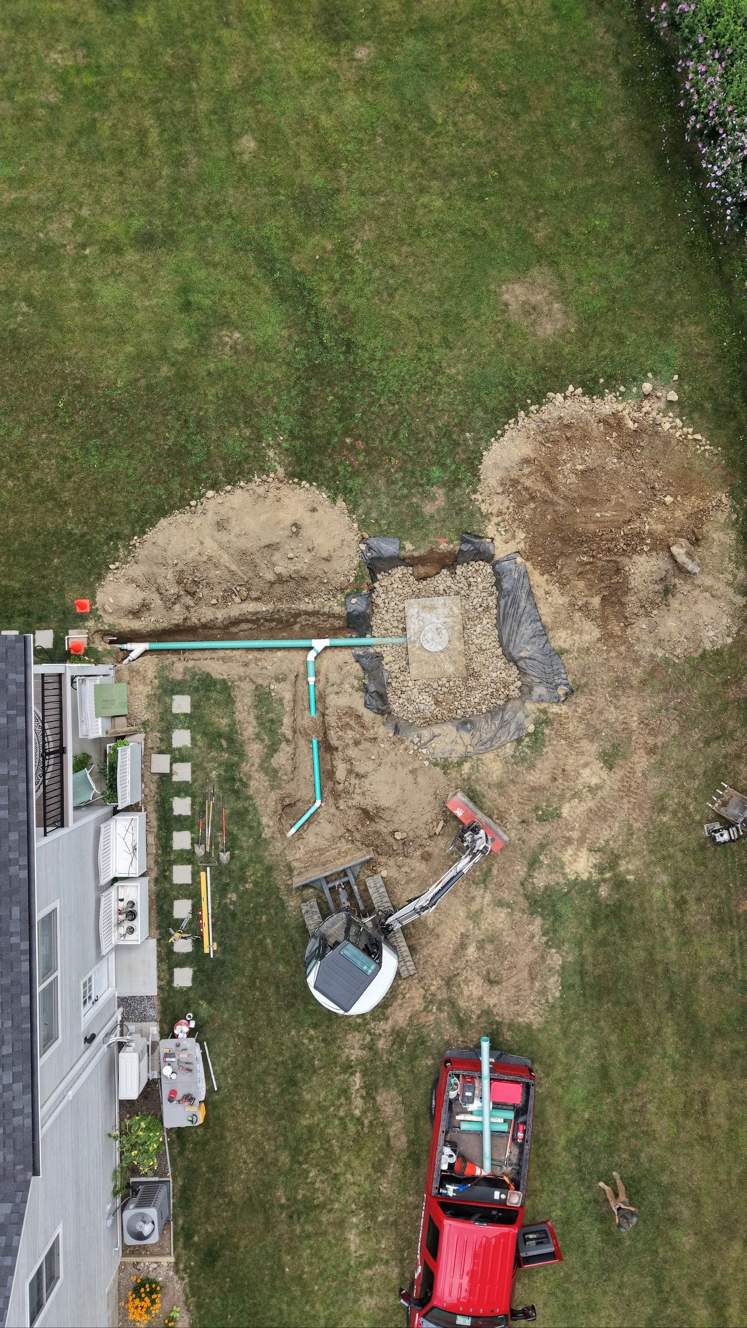 Aerial view of a residential backyard under construction with excavation and piping work, including a digger, trucks, and construction tools, next to a house with a patio, lawn, and flowering plants.