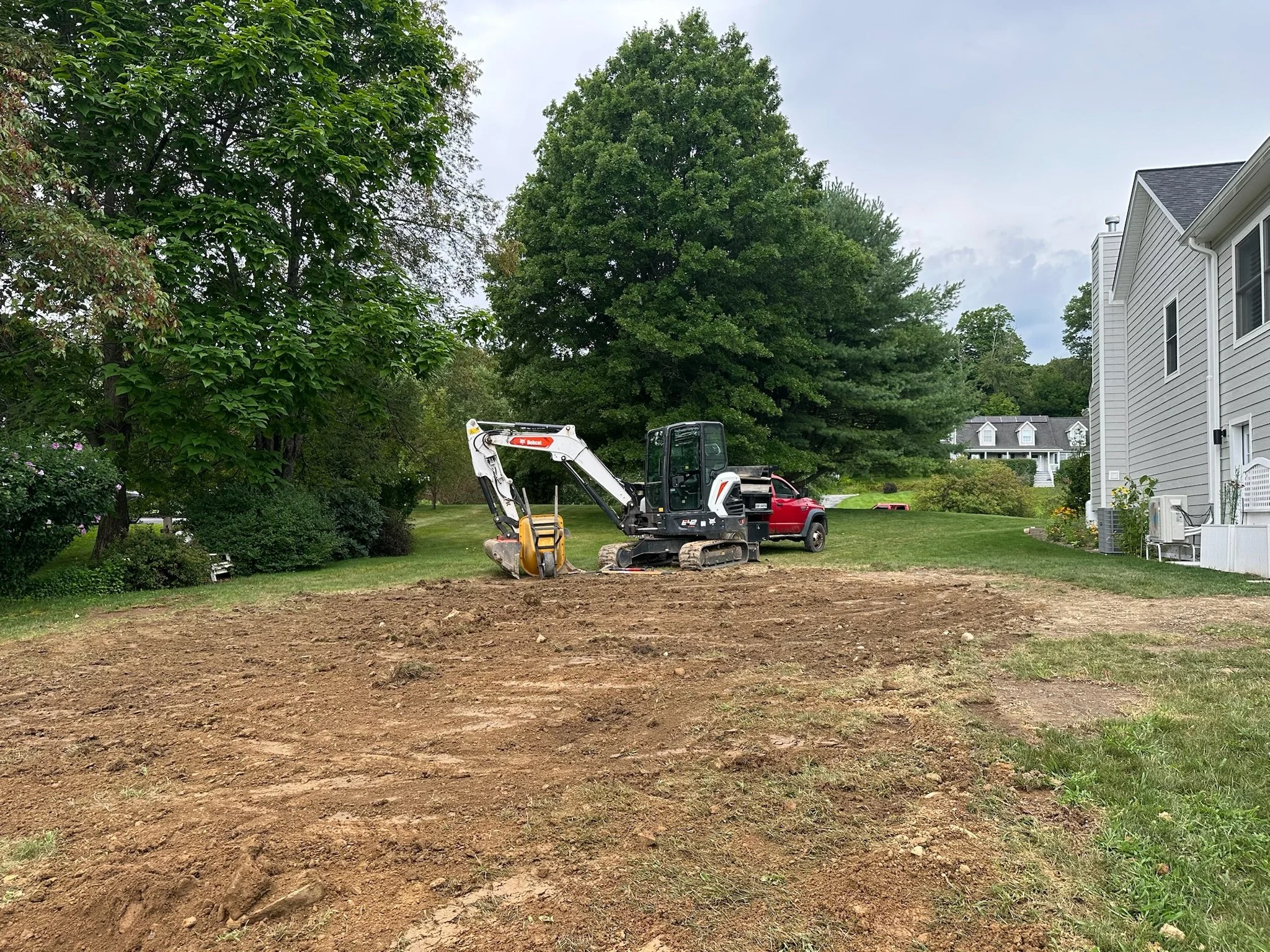 A backyard under construction with a compact excavator and a pickup truck parked nearby. The area has a mix of cleared soil and grass, with houses and trees surrounding the yard.