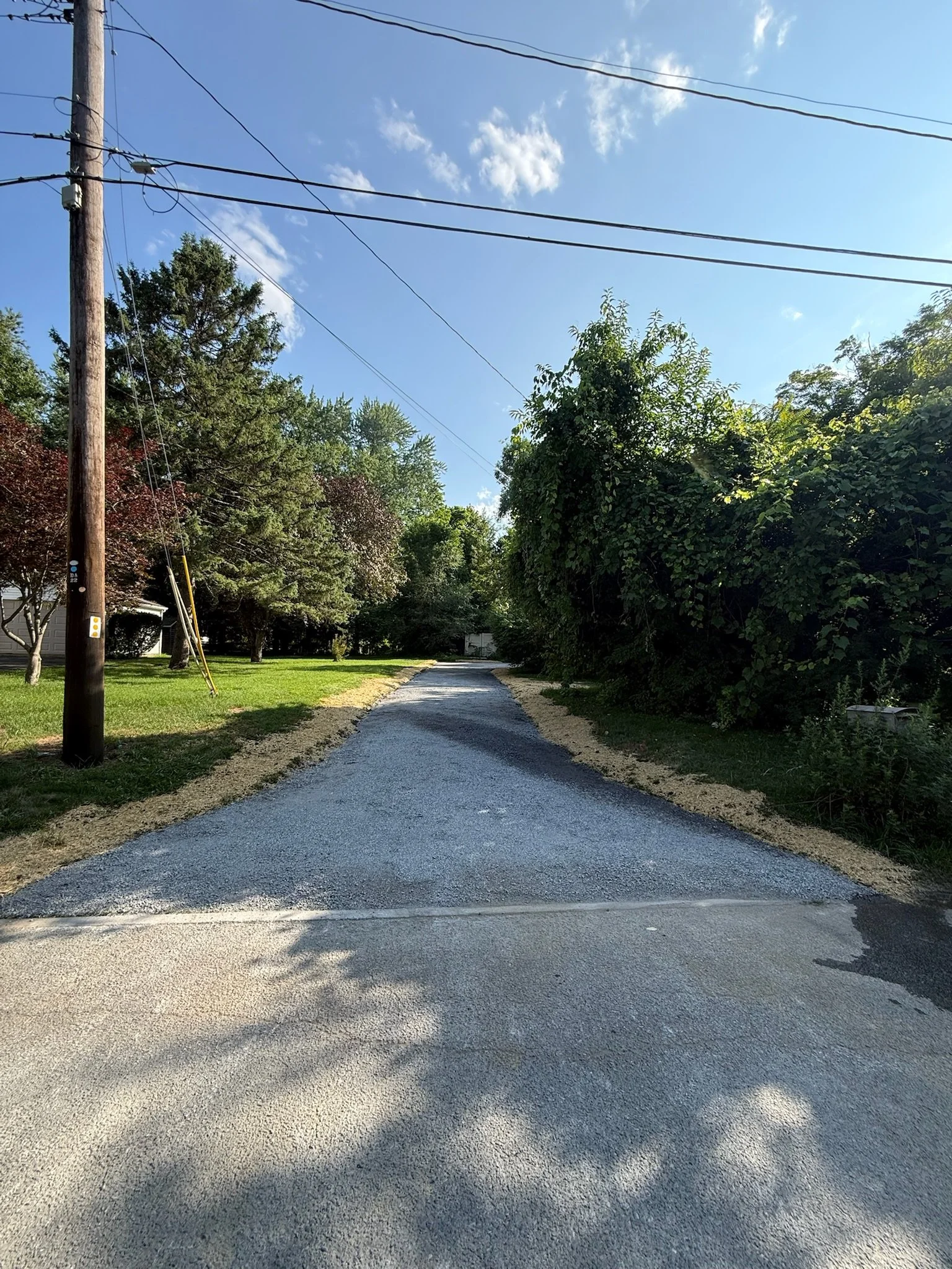 A gravel pathway in a residential area with lush green trees and bushes on both sides. Utility poles and wires are visible against a clear blue sky with some scattered clouds.