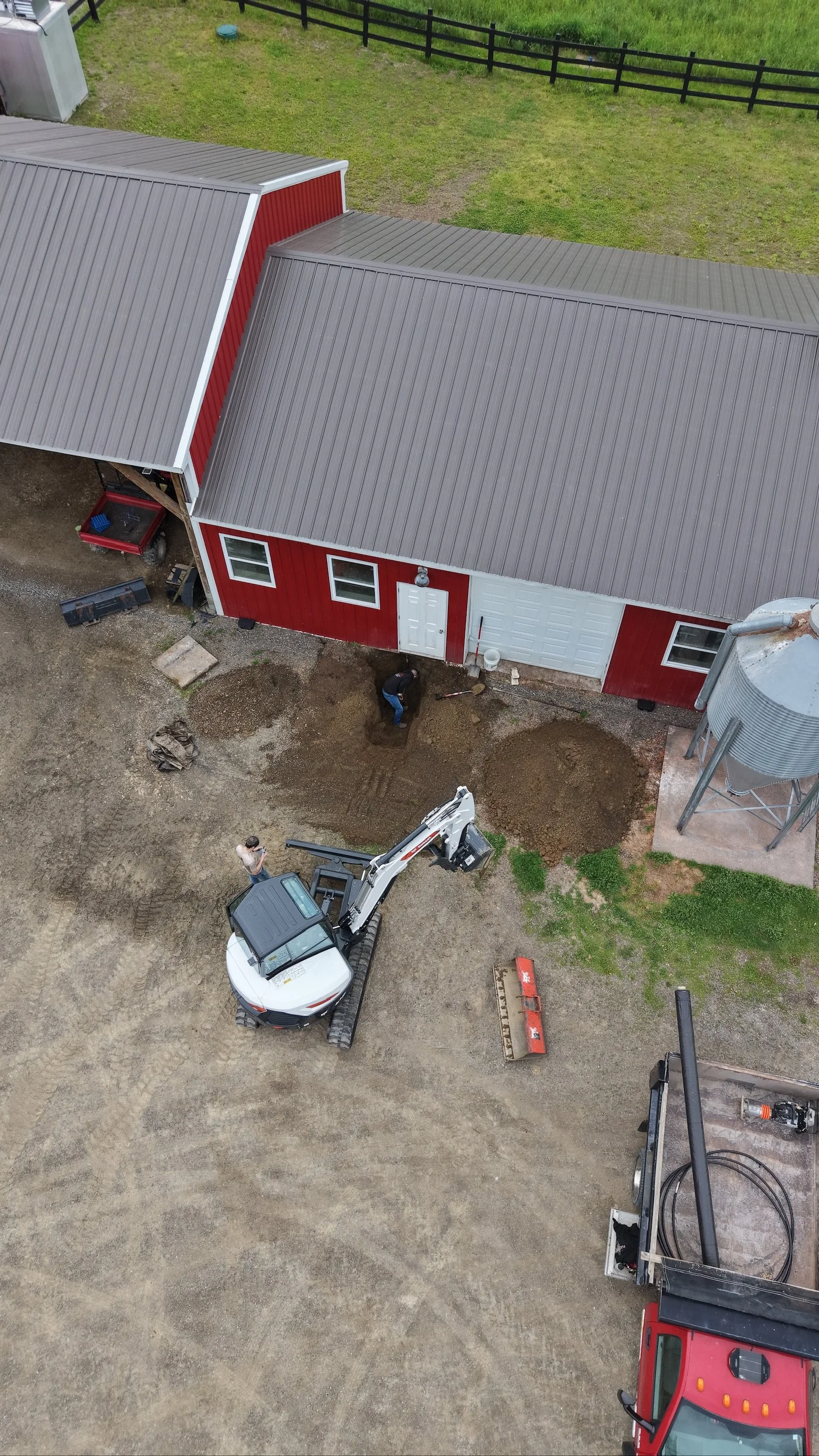 An aerial view of a farm building with a red exterior and a grey metal roof. Two people are working on the ground in front of the building, with construction equipment and vehicles nearby. There are patches of dirt and gravel, a gravel driveway, and 