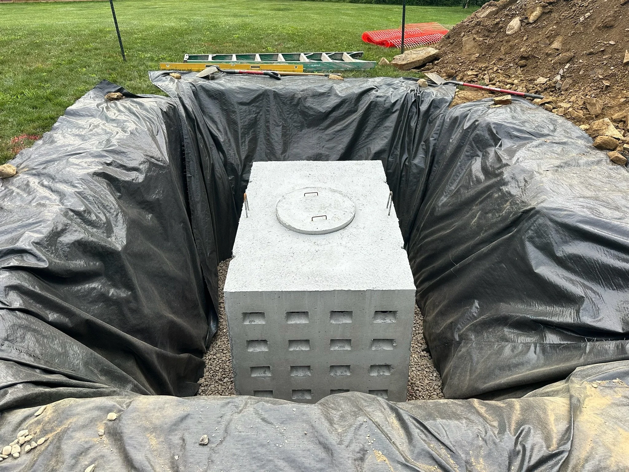 A construction area with black plastic sheeting, a cinder block, and tools including a level, a crowbar, and a rake, beside a mound of dirt.