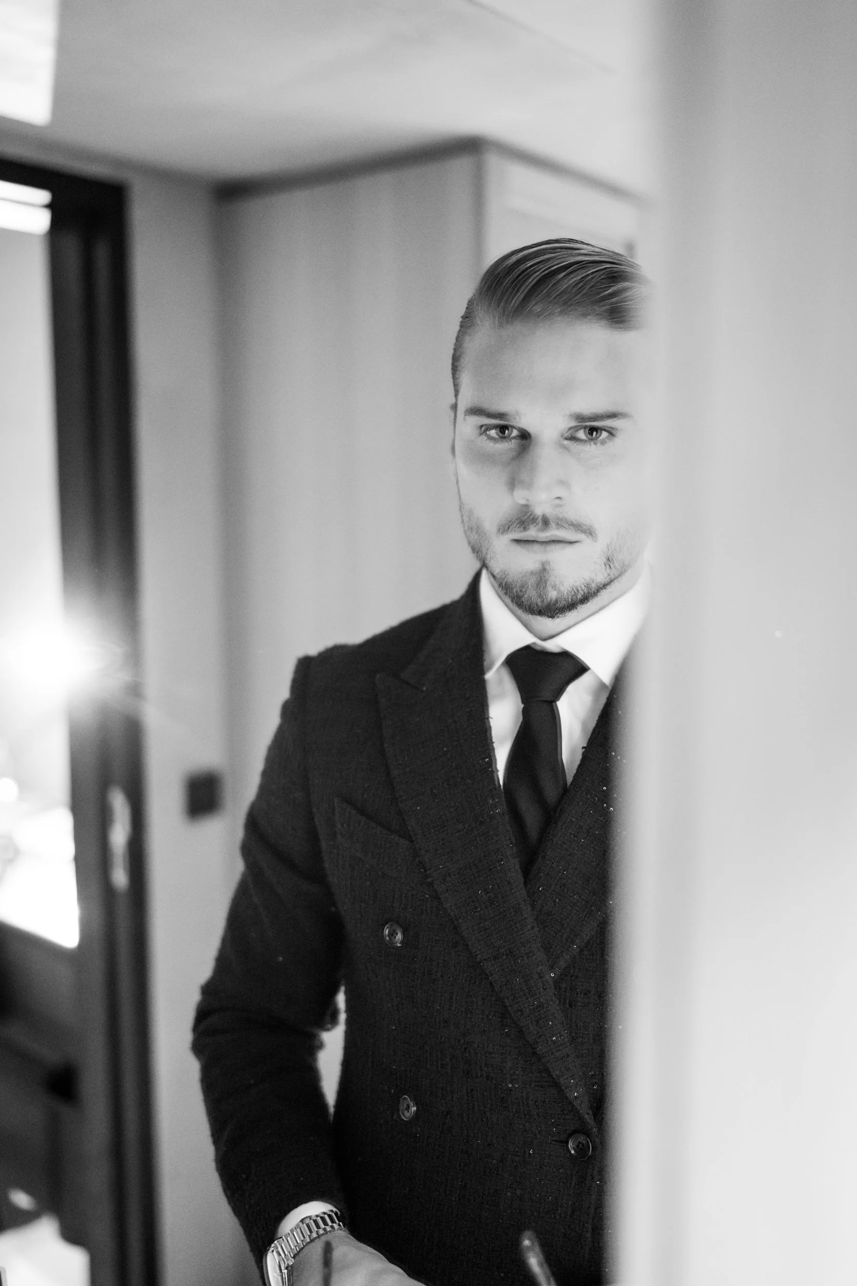 Schwarz-weiße Fotografie eines jungen Mannes (Rurik Gislason)  in formeller Kleidung, der sich im Gesicht im Spiegel betrachtet, mit glatter Haarfrisur und Stilvoller Kleidung, in einem modernen Raum.