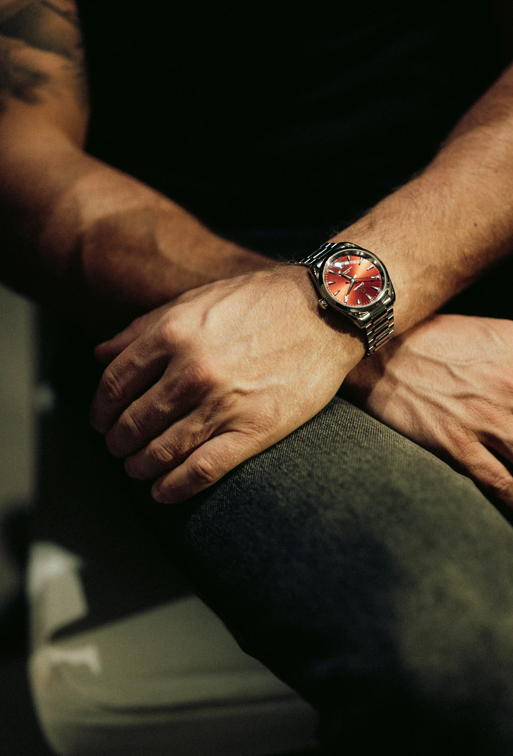Nahaufnahme eines Mannes (Rurik Gislason) mit einer Metallarmbanduhr mit rotem Zifferblatt am Handgelenk, sitzend mit verschränkten Armen.