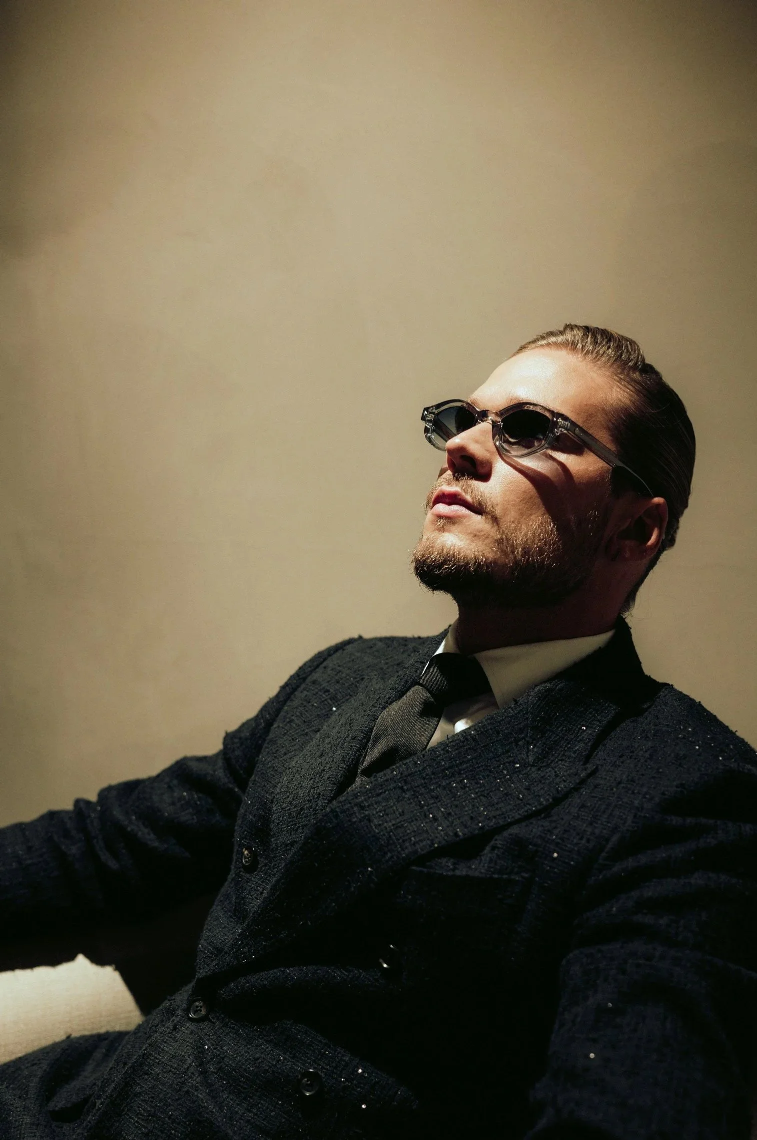 Ein Mann (Rurik Gislason) mit Bart, Sonnenbrille, schwarzem Anzug und Krawatte sitzt entspannt gegen eine beige Wand.