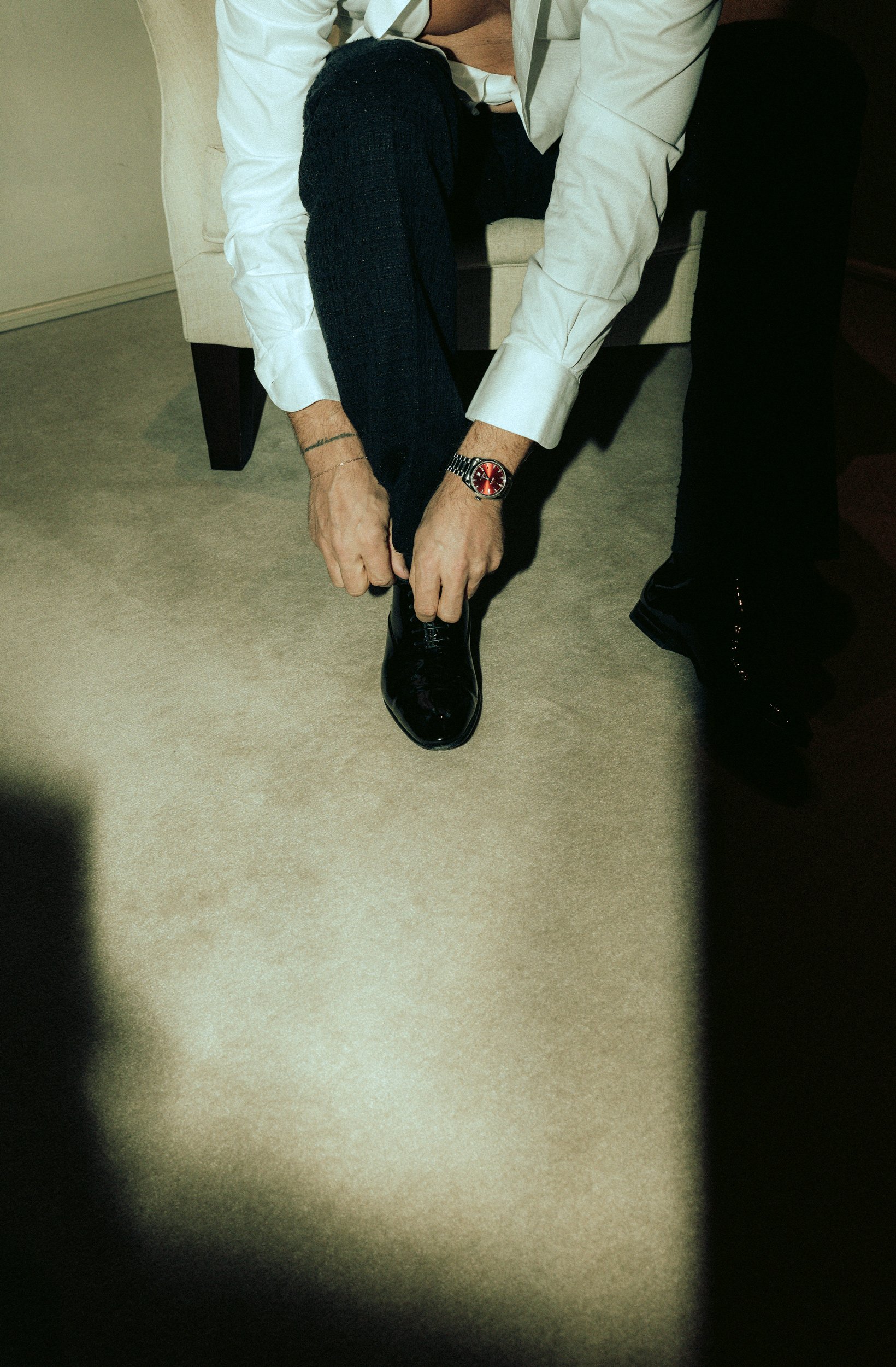 Ein Mann  (Rurik Gislason) zieht einen schwarzen Schuh an, trägt weiße Hemd, schwarze Hose, Armbanduhr, sitzt auf einem Stuhl, auf Teppichboden, im Raum.