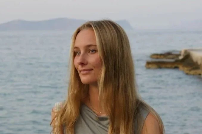 En ung kvinna med långt blont hår står vid havet.