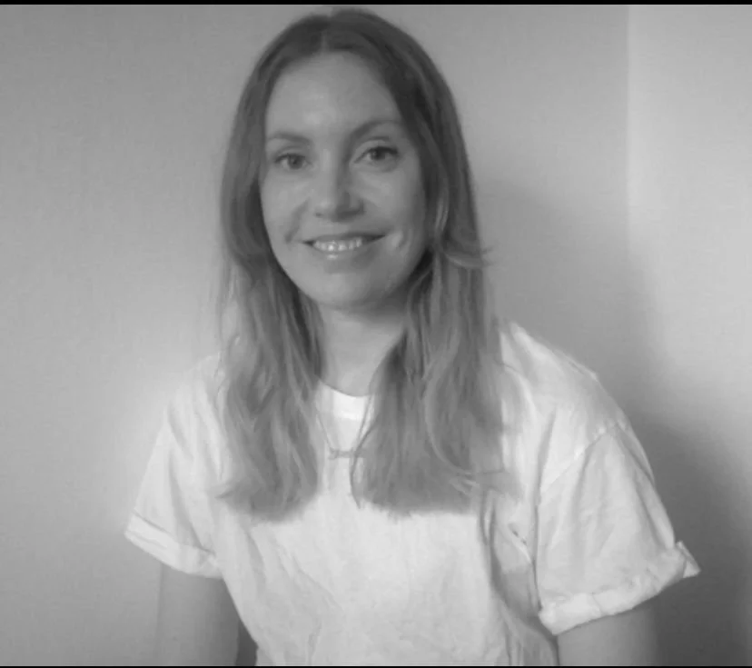 Kvinna med långt, vilt, lockigt hår, ler mot kameran, bär vit t-shirt, står mot vägg, svartvitt foto.