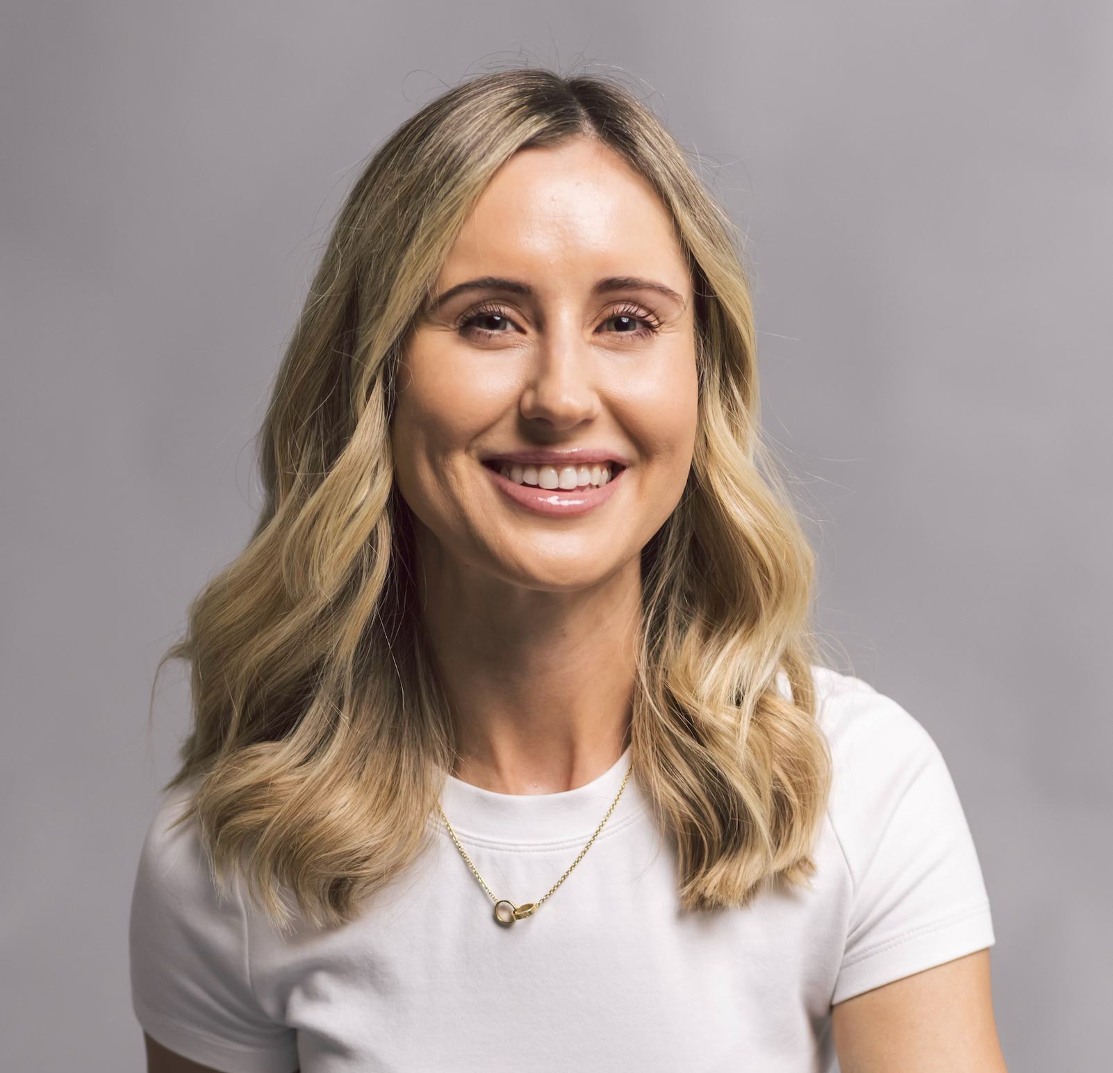 blonde woman is smiling at the camera. She is wearing a white shirt, a gold necklace. And she has kind eyes.