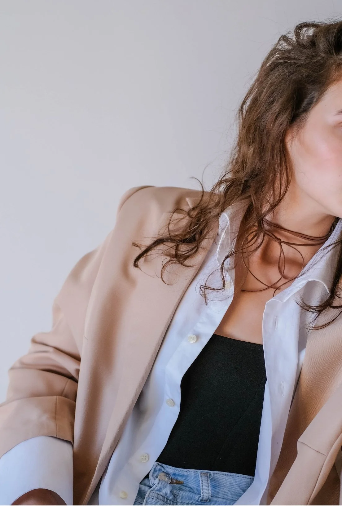 woman is sitting. wearing a beige blazer, a white button up shirt that is open, showing her black undershirt tucked into jeans