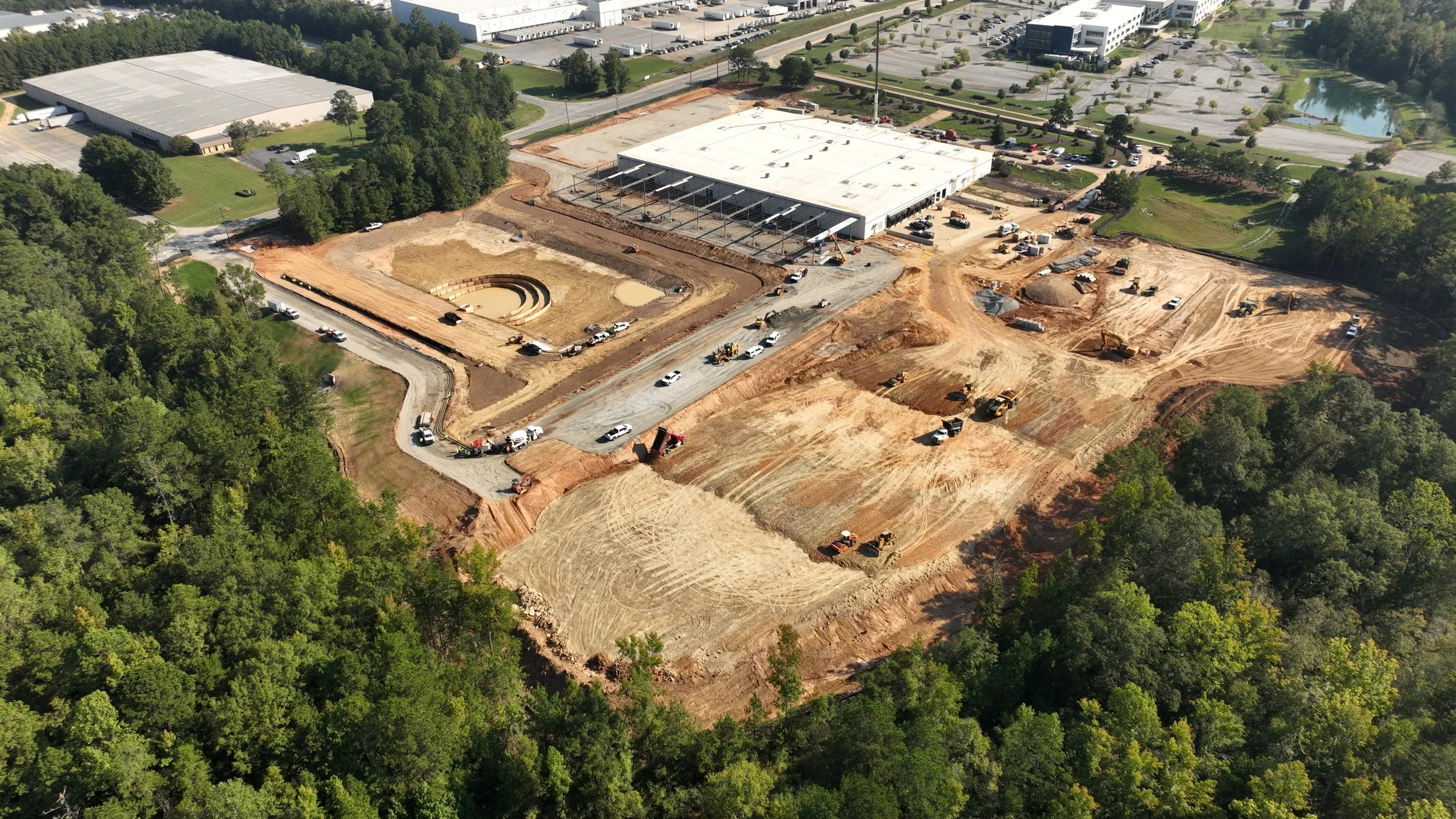 Wide aerial view of a construction site showing the main building, loading canopy under construction, site basin, access drive, and surrounding earthwork.