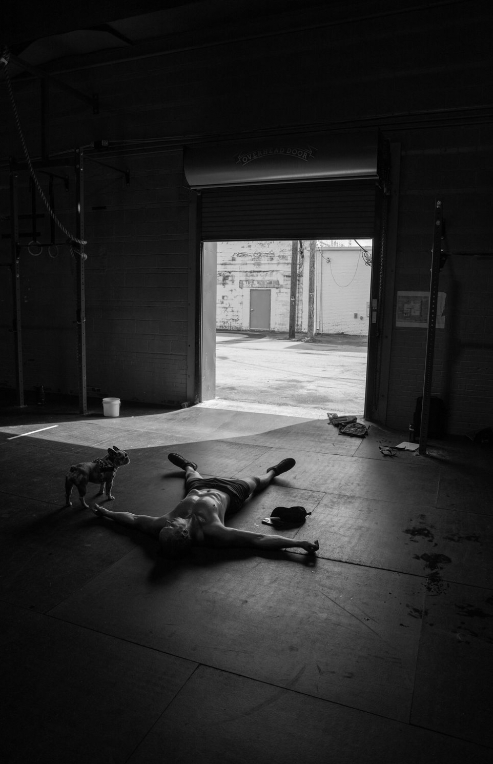 A shirtless man lying on the floor of a gym with arms and legs outstretched, a small dog near him, and gym equipment visible in a garage-style setting, with sunlight coming through an open door to the outside.