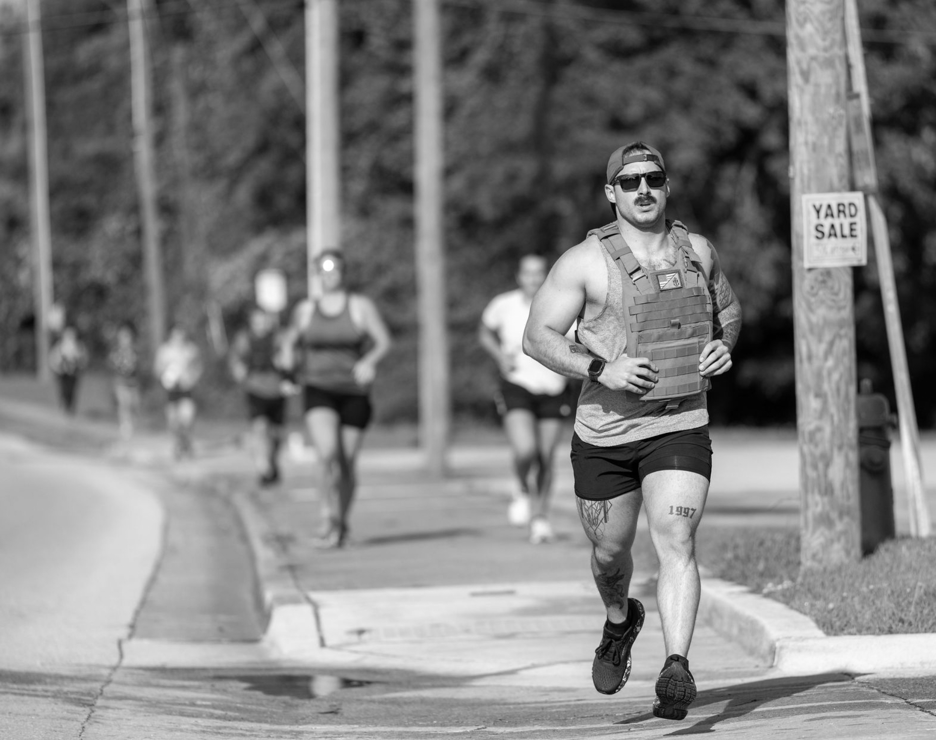 A man running along a sidewalk, wearing sunglasses, a cap, a sleeveless shirt, shorts, and a hydration vest, with a tattoo on his leg and arm, and blurry runners in the background.