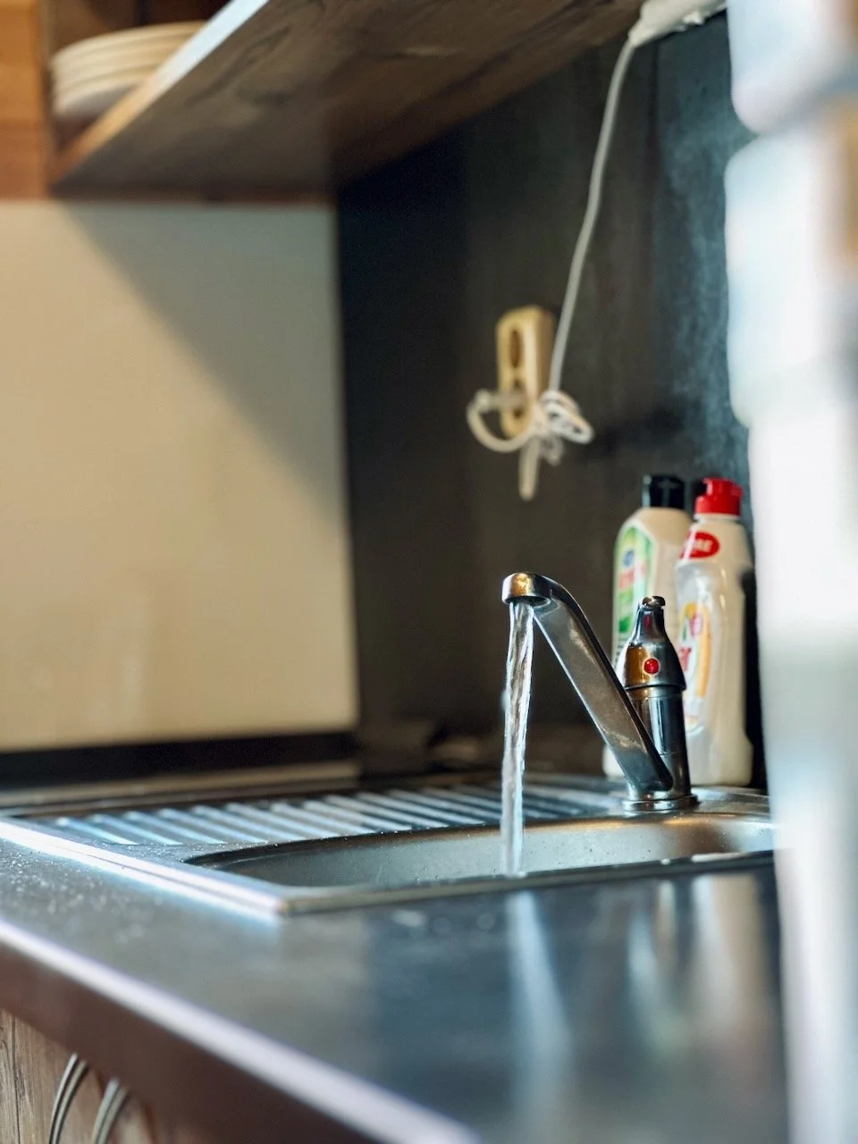 Kitchen sink with running water, dish soap bottles on the counter, wooden shelf above, and wall outlet with an unplugged cord.